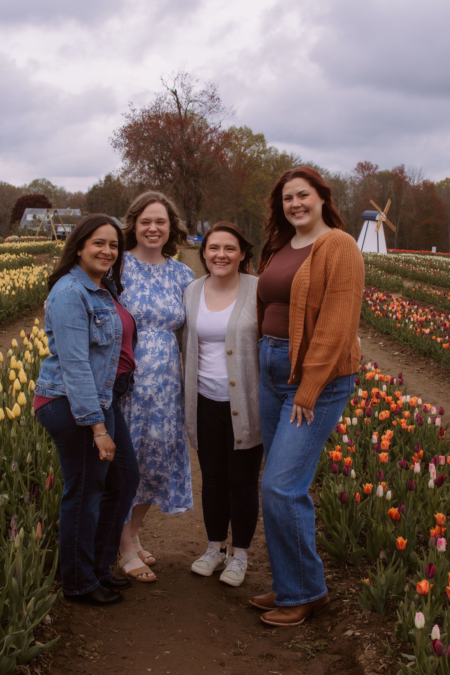 Four women smiling together in a colorful tulip field with a windmill in the background on a cloudy spring day.
