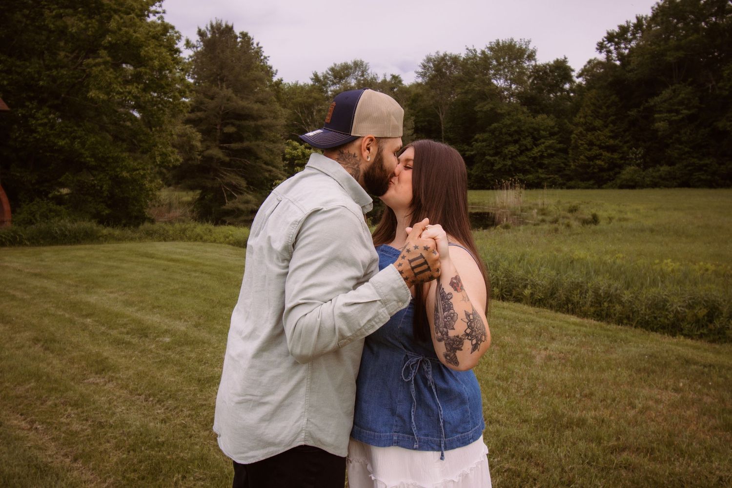 A couple shares a romantic kiss outdoors in a lush green field, woman wearing denim top with floral tattoo on arm.