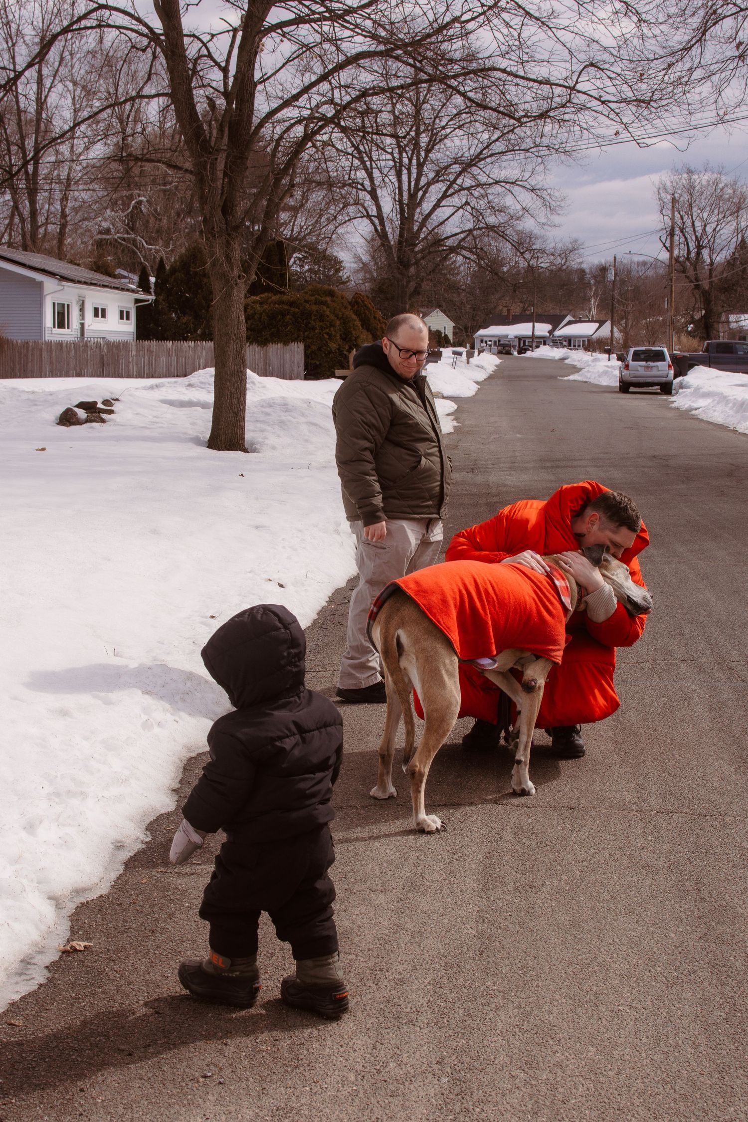 Family with toddler and large dog in orange coat on snowy suburban street in winter.
