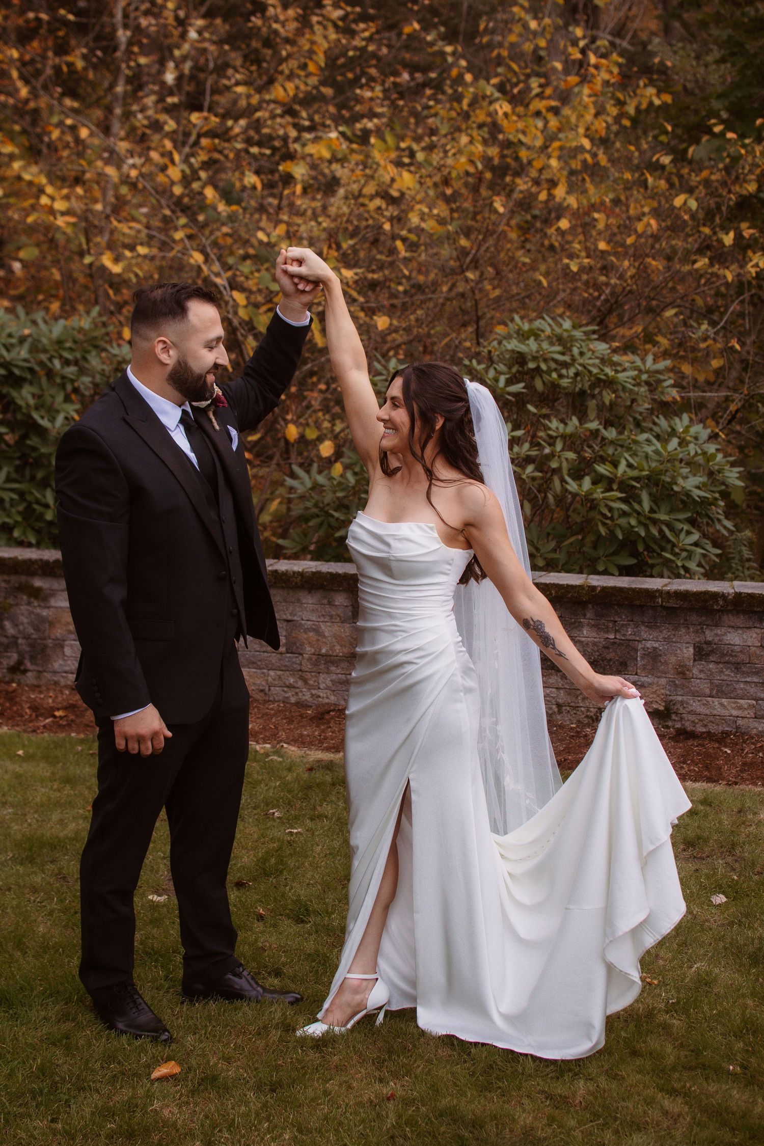 A couple in wedding attire dances together on grass surrounded by autumn foliage.