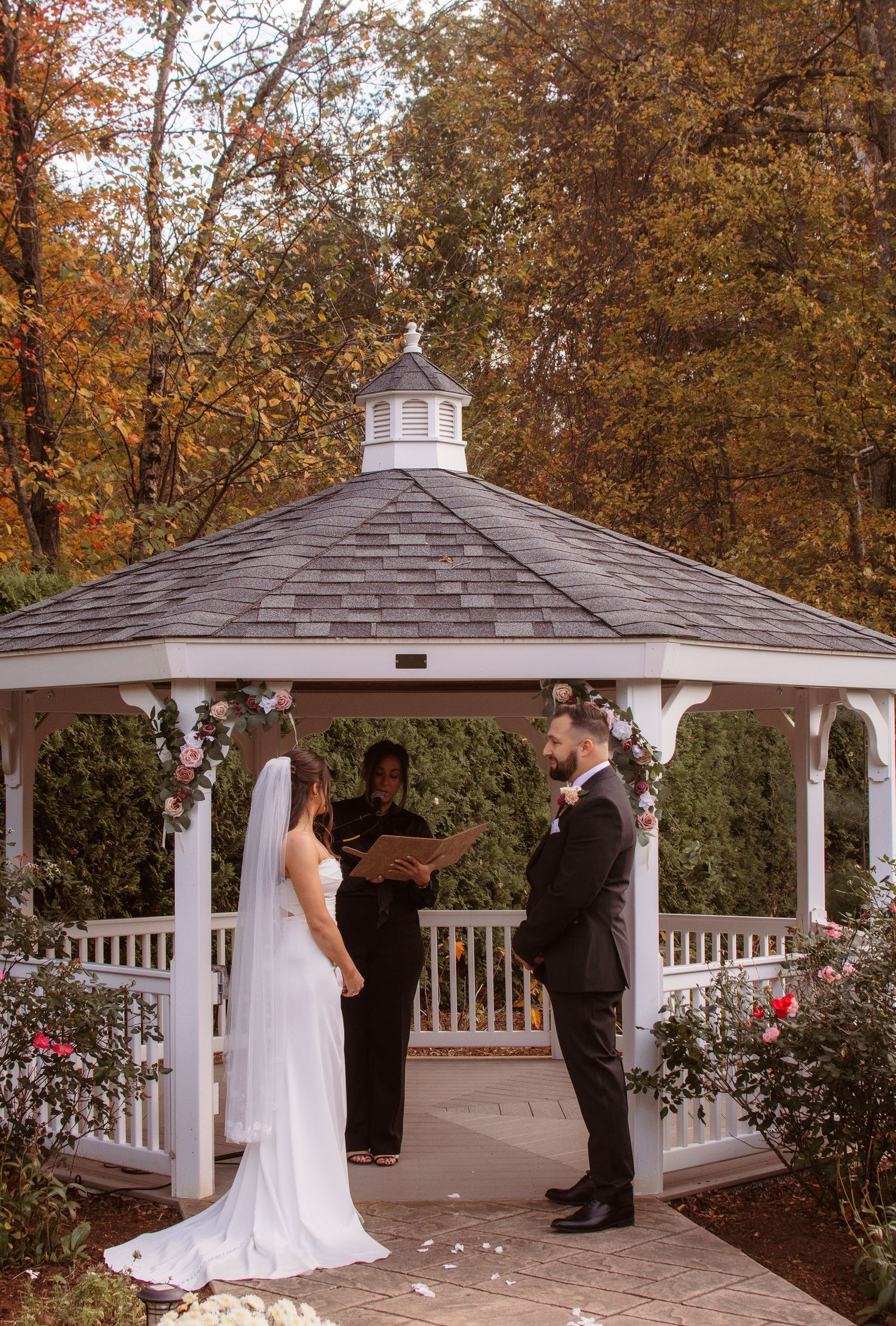 A couple shares a romantic moment under a white gazebo surrounded by autumn foliage.