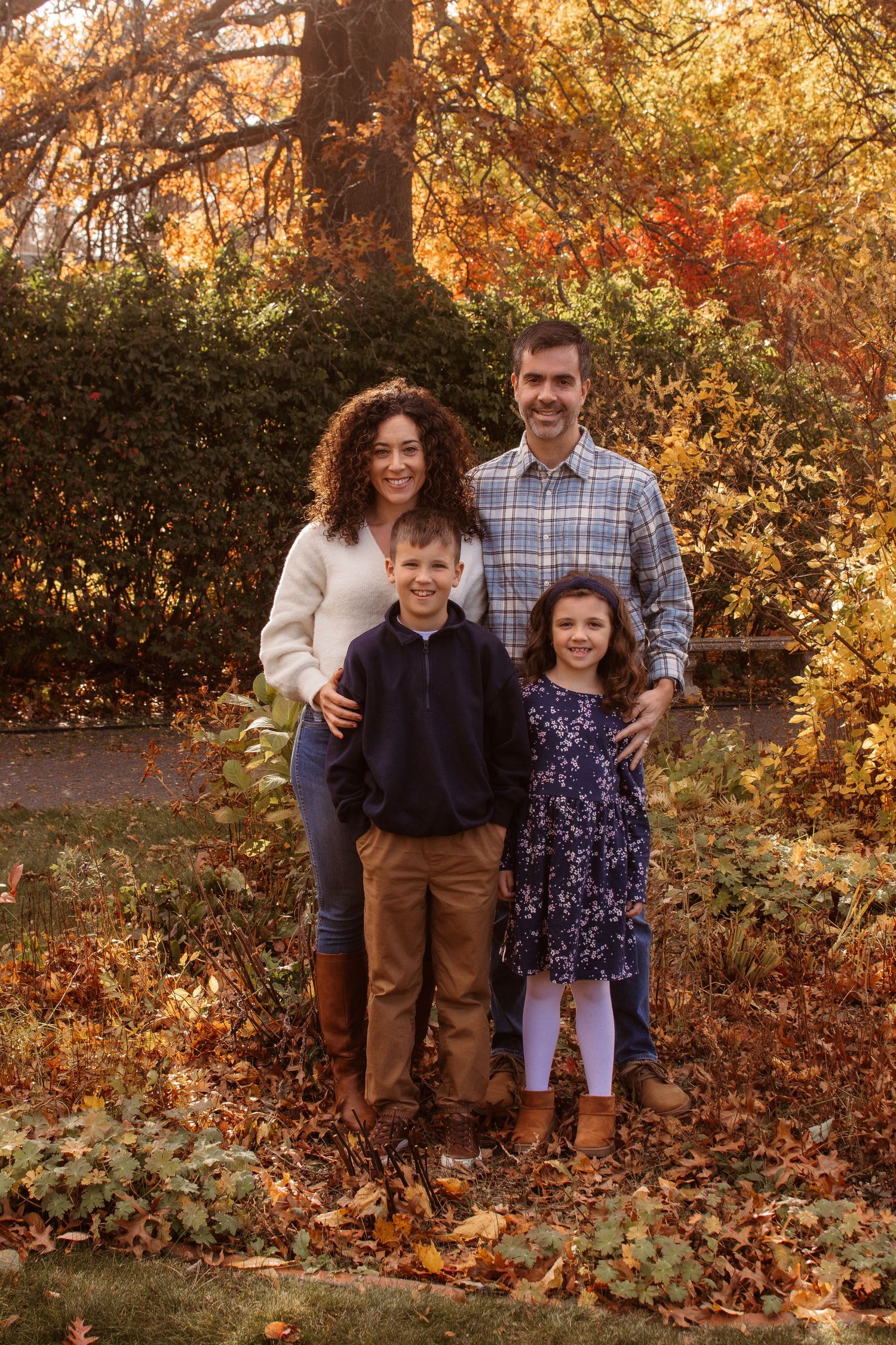A family poses together outdoors during autumn with golden foliage and fallen leaves in the background.