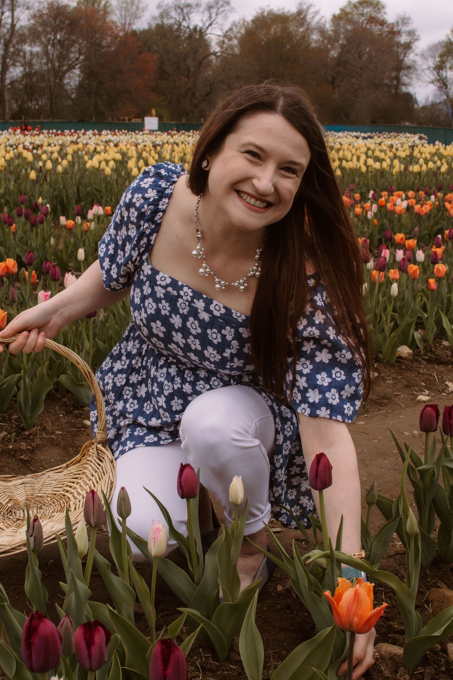 Smiling woman in blue floral top and white pants crouches beside tulips in a vibrant flower field.