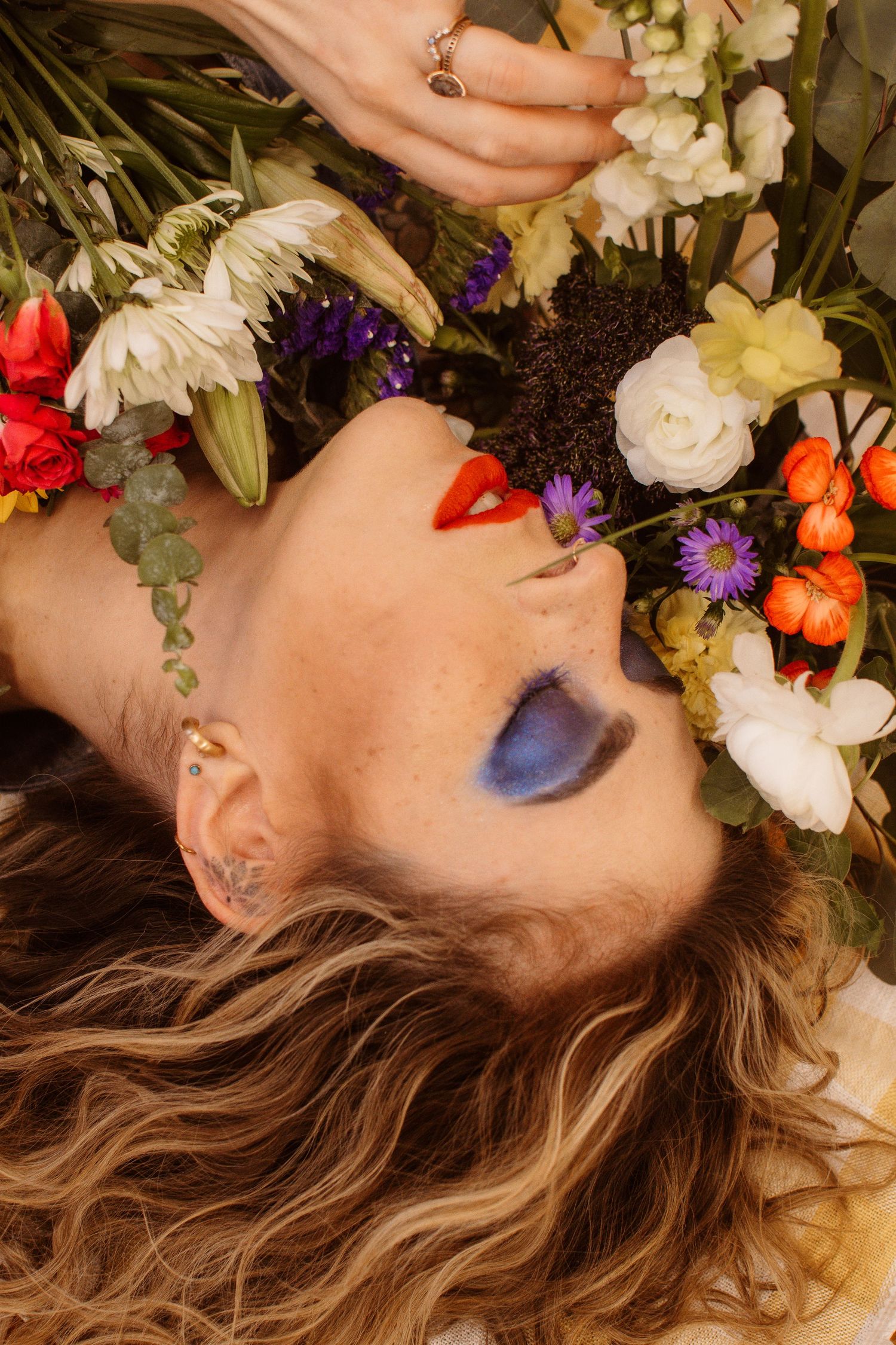 Woman lying among colorful flowers with bold blue eyeshadow, red lips, ear piercings, and flowing hair.