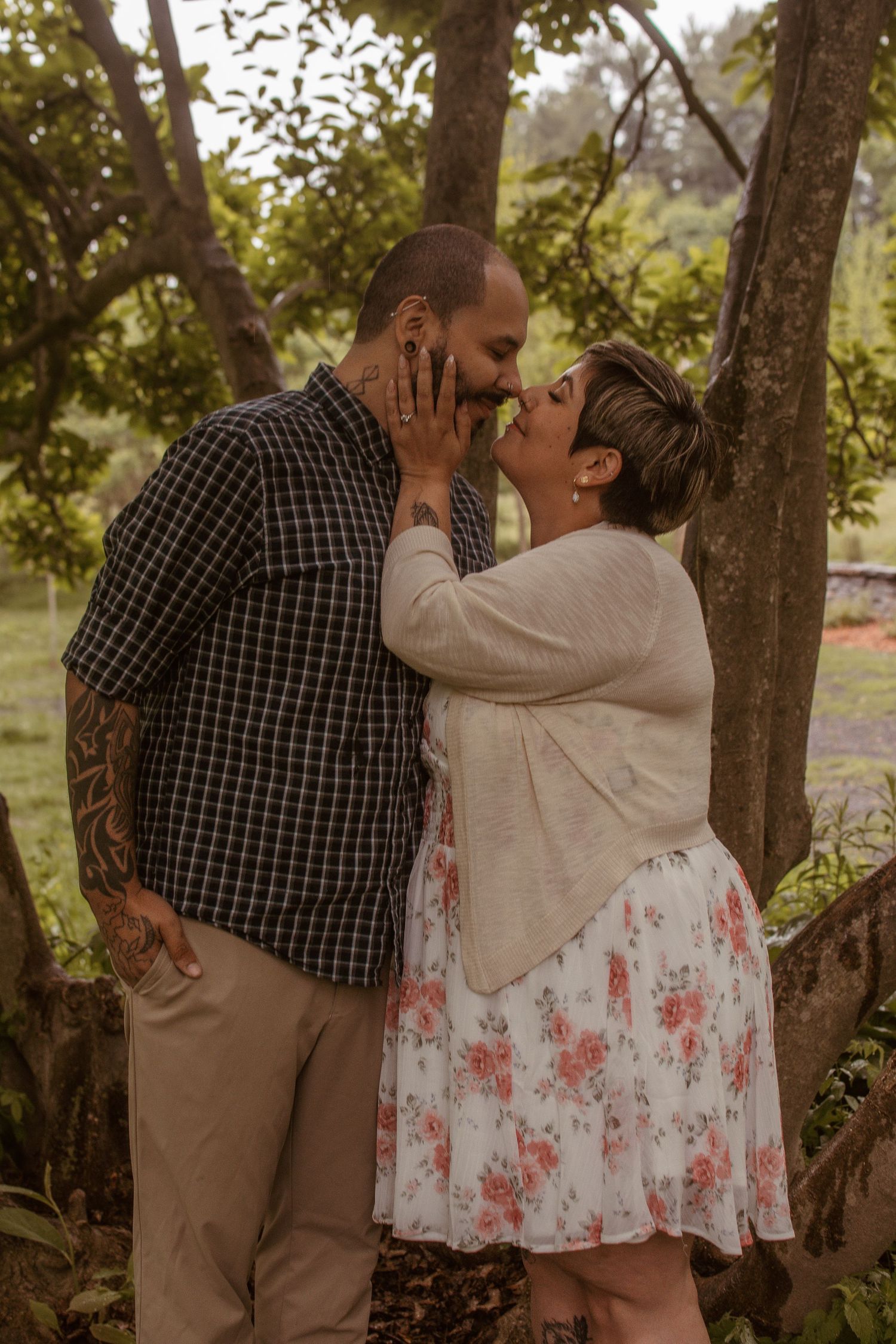 Couple sharing a romantic kiss outdoors, surrounded by lush green trees, man with tattoos in plaid shirt, woman in floral dress.