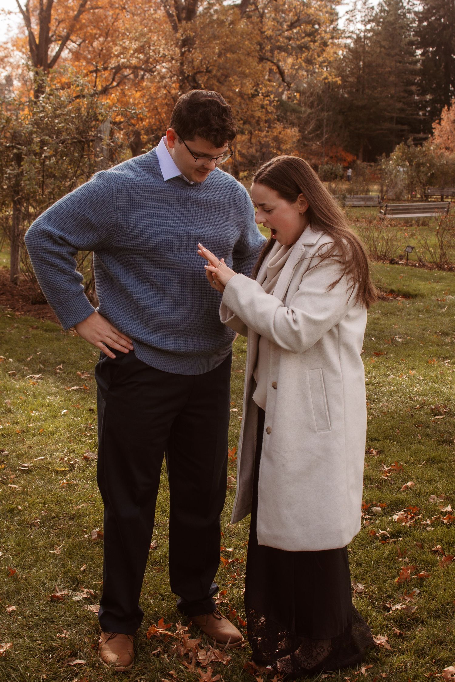 A couple stands together on an autumn lawn, with one person showing something on their phone while wearing a white coat.