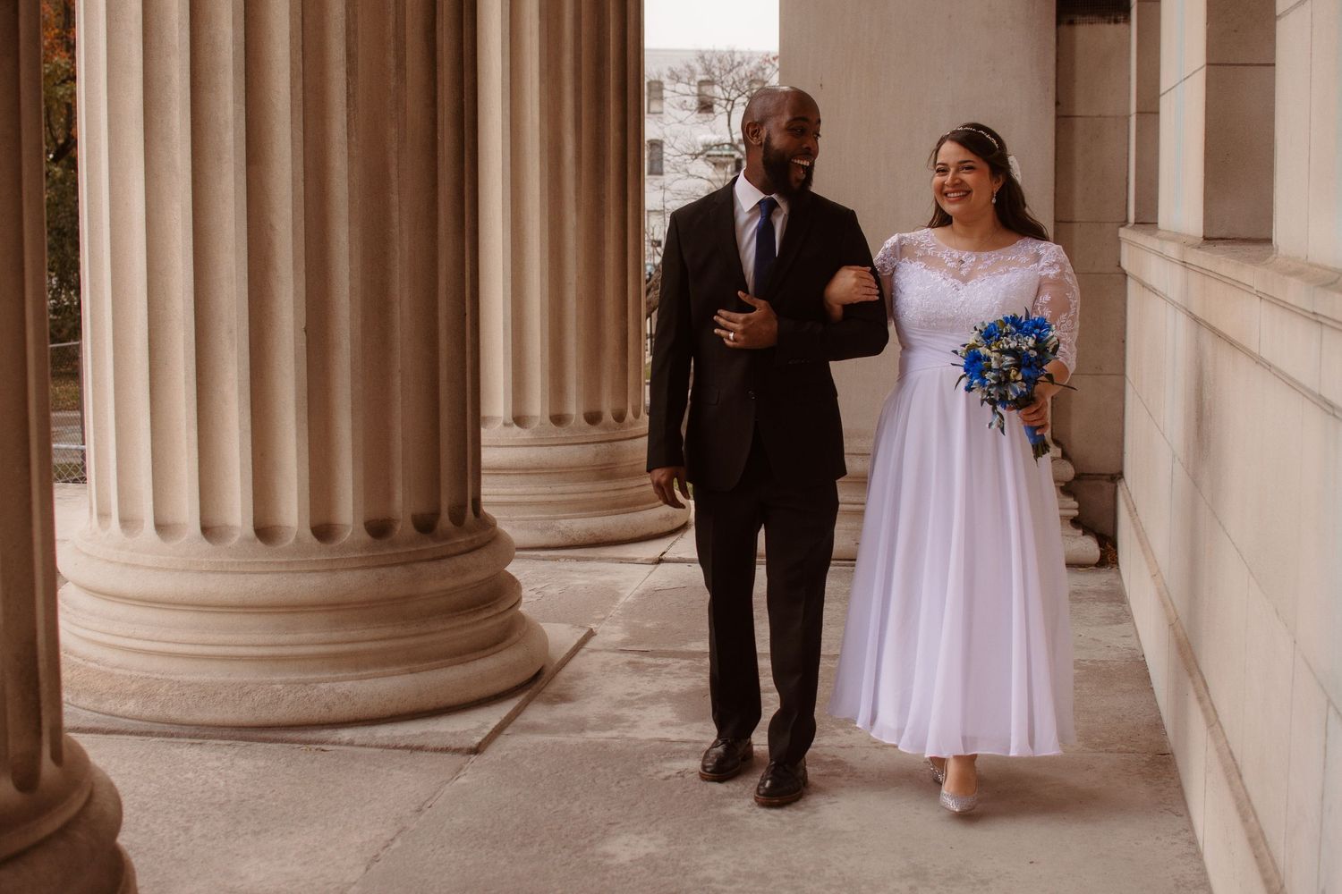 A couple walks through an elegant colonnade hallway with white pillars and stone architecture.