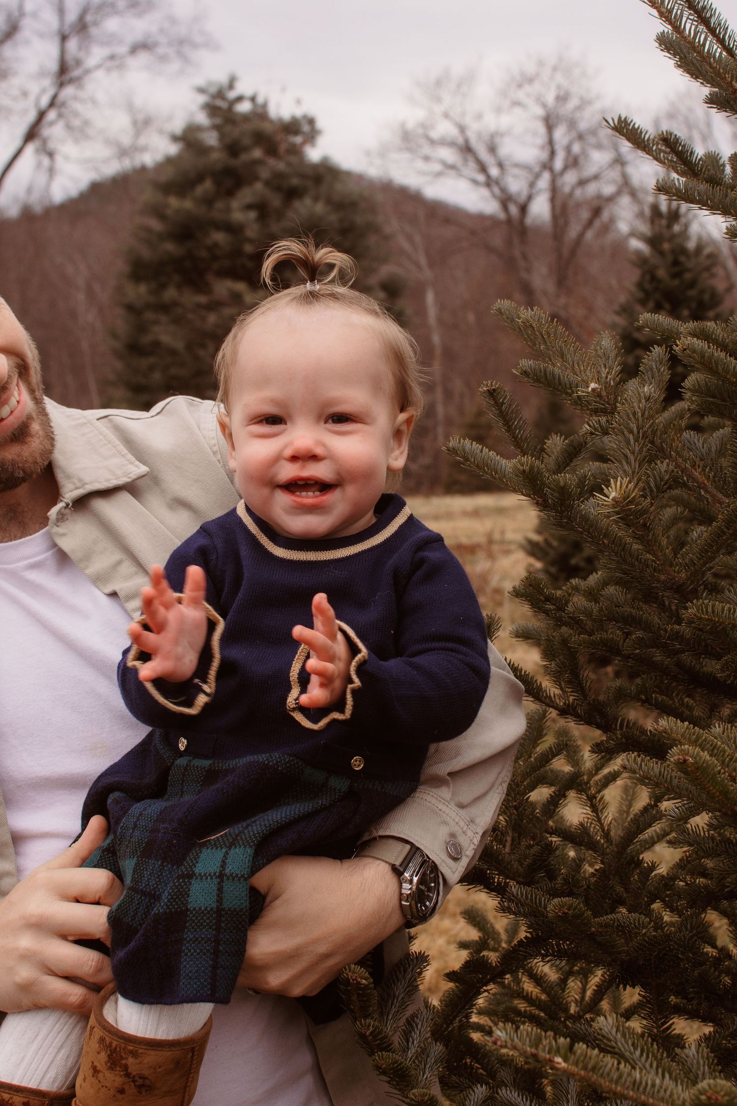 A series of five photos showing a smiling baby in a navy sweater and plaid pants being held near evergreen trees.