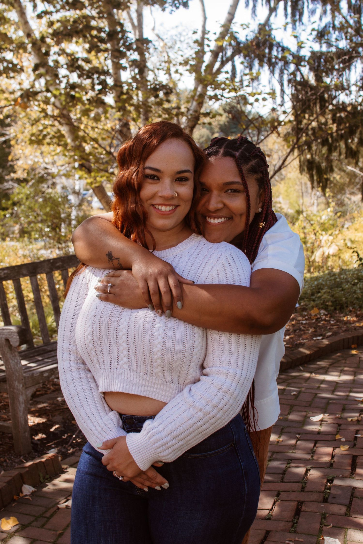 Two people share a warm embrace while sitting on brick steps in a sunlit outdoor setting with trees in the background.