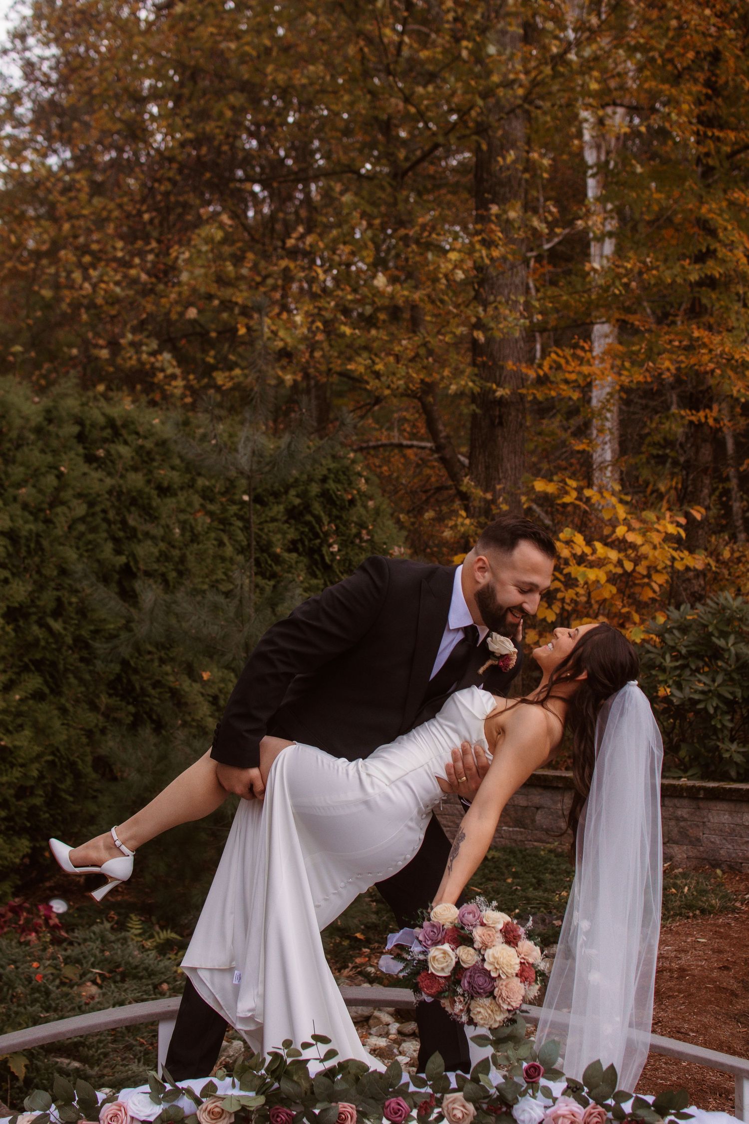 Romantic wedding couple dips and poses near floral arrangements with fall foliage backdrop.