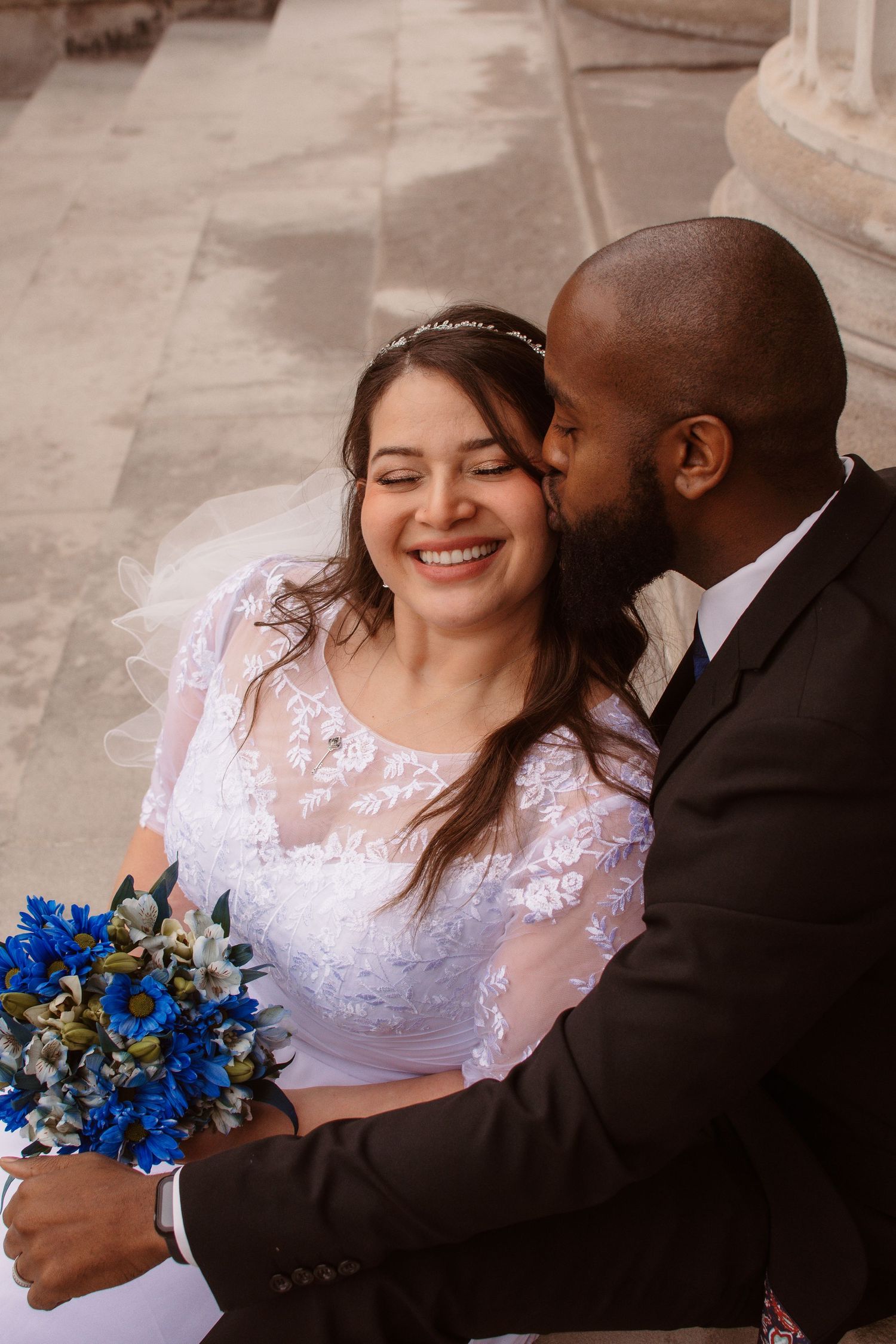 A joyful wedding couple embraces while holding a vibrant blue bouquet, the bride wearing a lace dress with long sleeves.