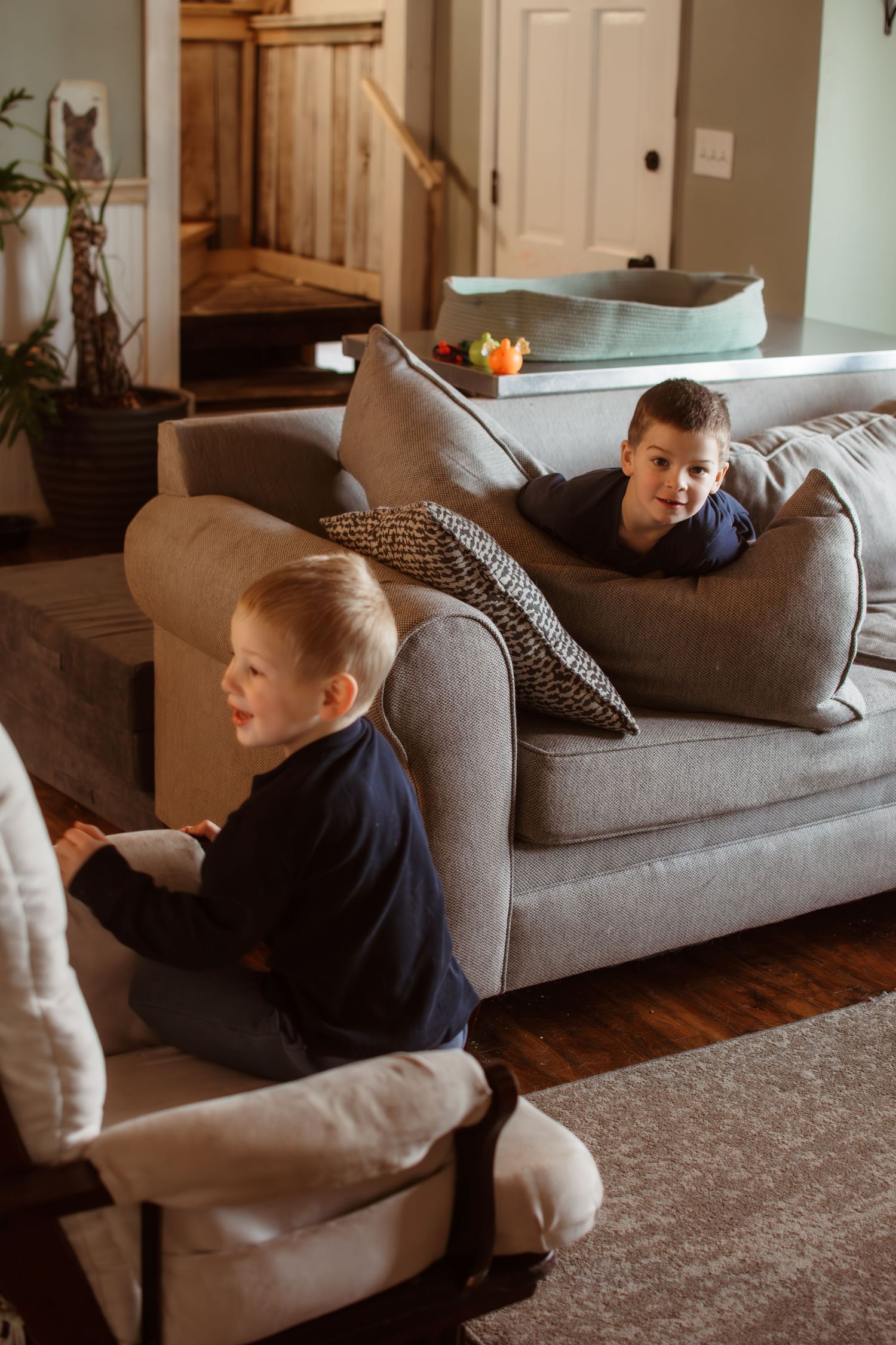 Two young ones relaxing on a grey couch in a cozy living room with hardwood floors and warm lighting.