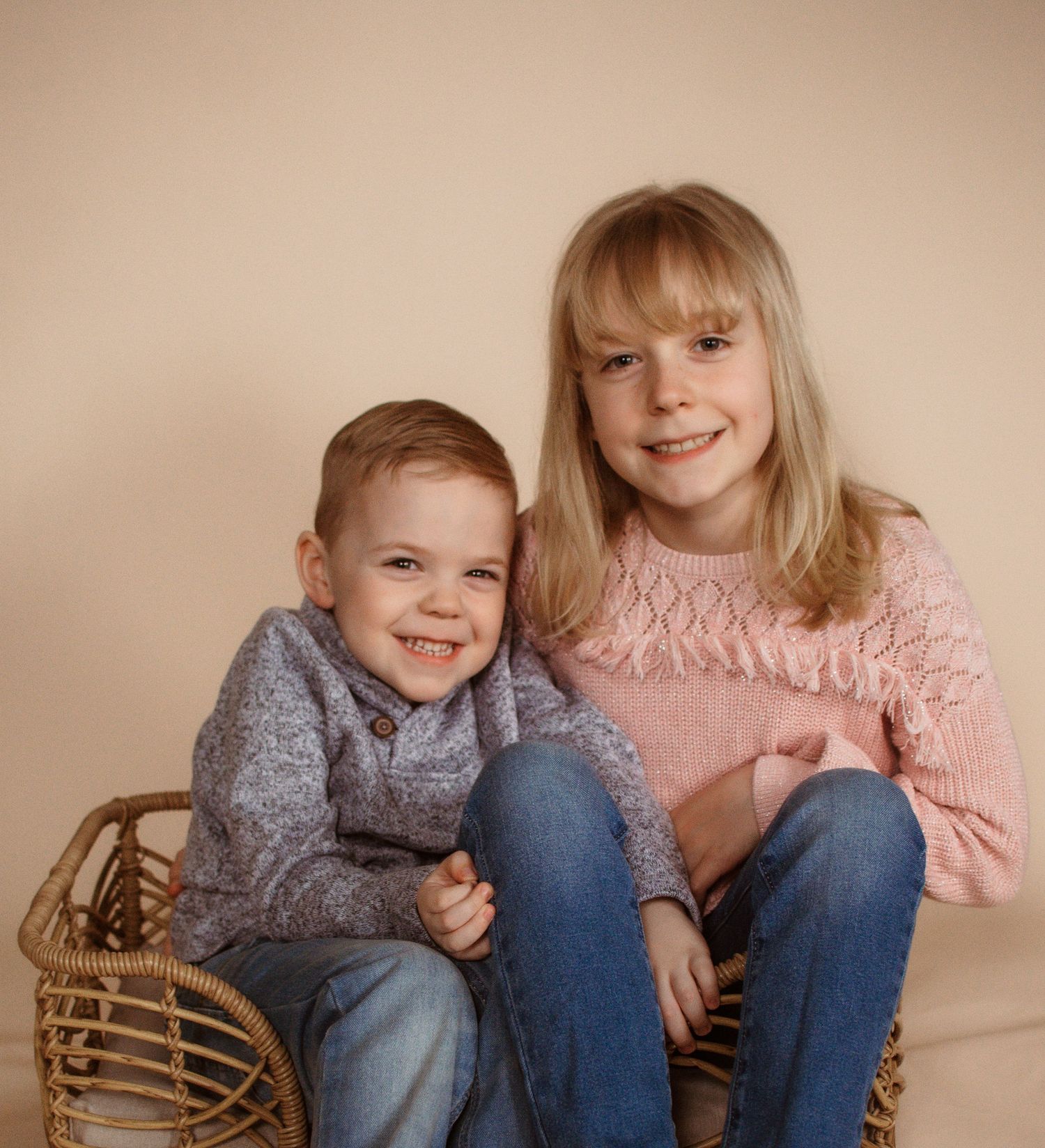 Two young children in cozy sweaters sit together smiling on a beige background with a woven basket beside them.