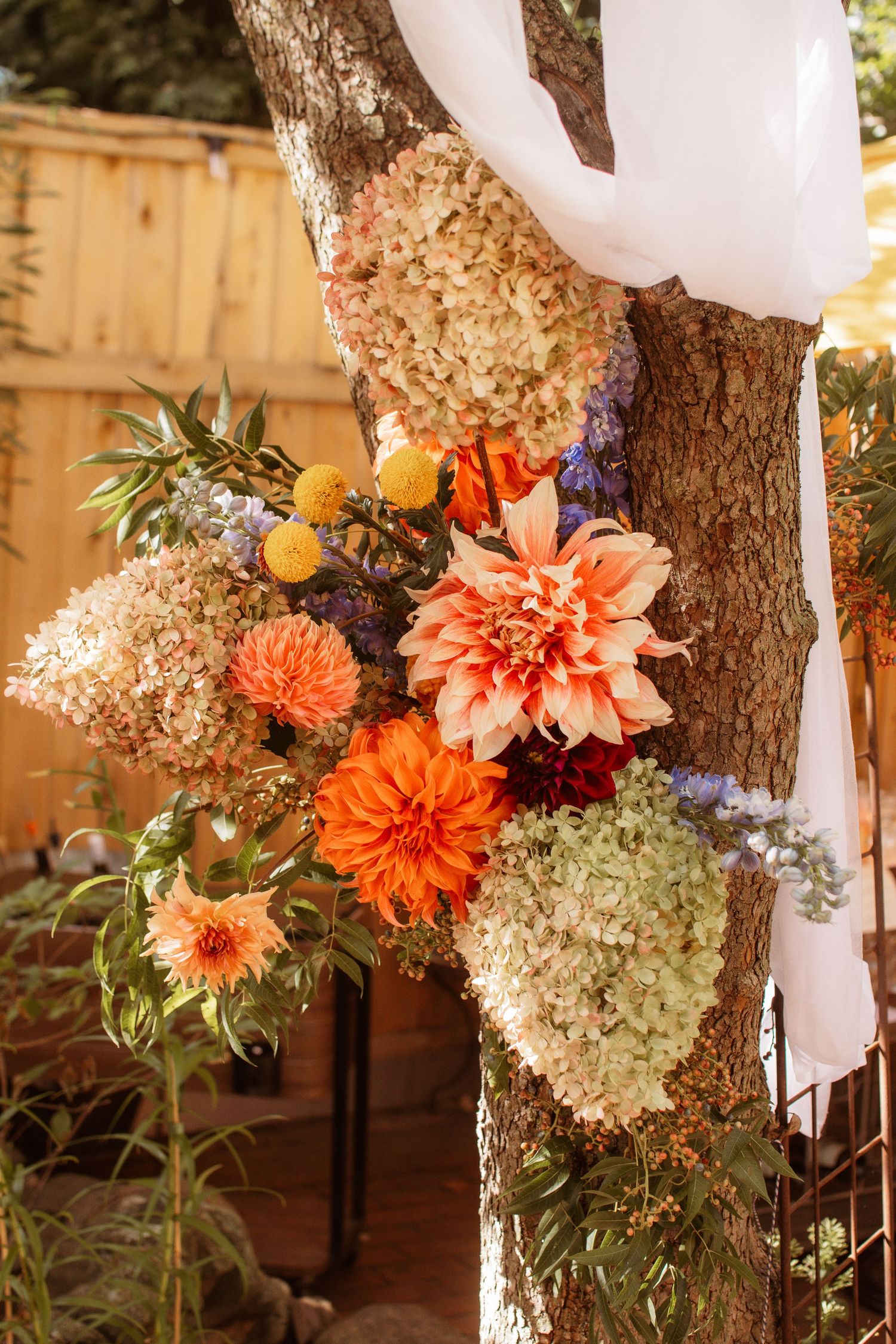 Vibrant orange dahlias and white hydrangeas arranged on a tree trunk with flowing white fabric draping.