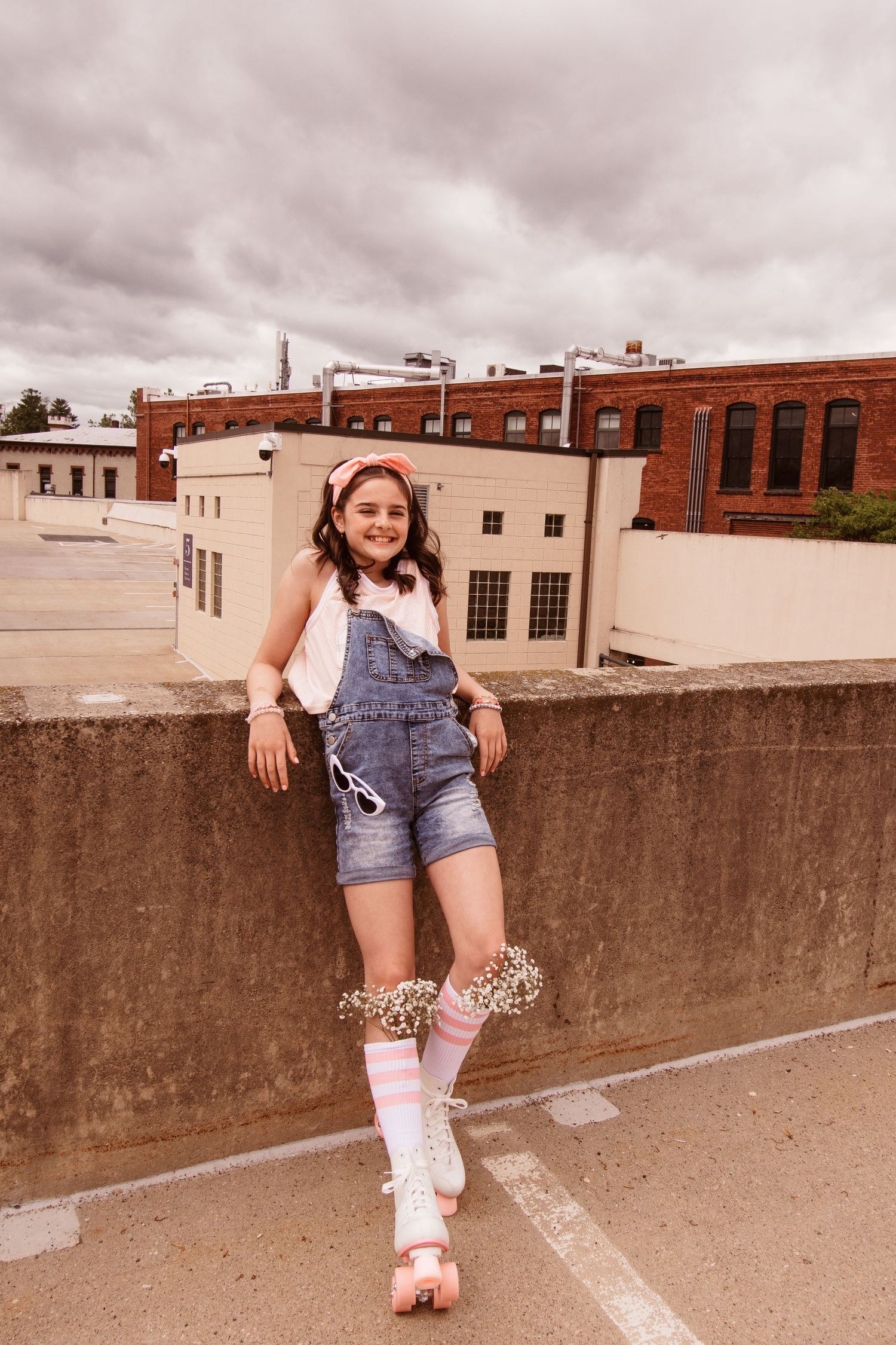A person wearing denim overalls and white boots poses on a concrete ledge in an urban setting while shot in a vintage style.