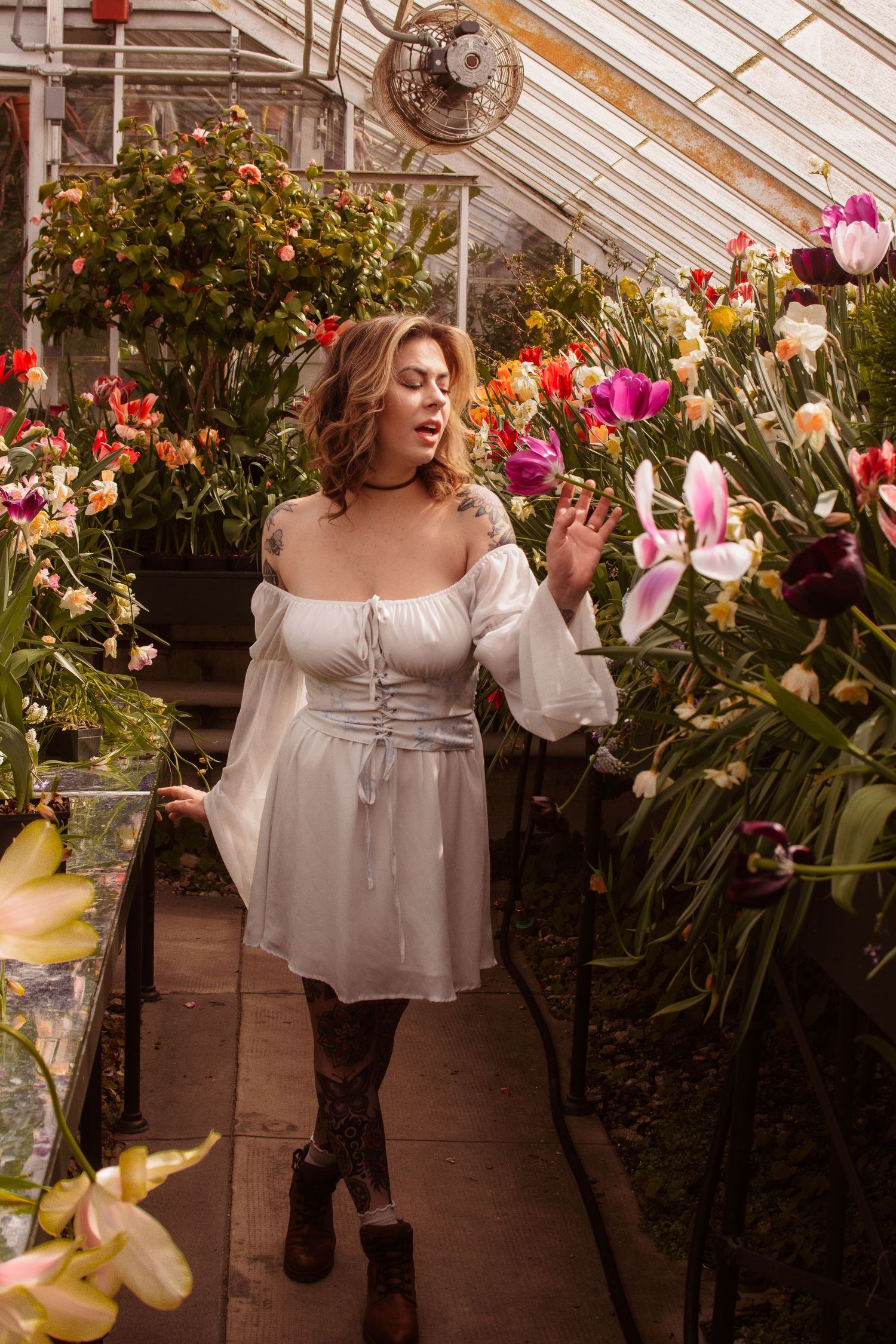 Woman in white off-shoulder mini dress poses among colorful flowers in a lush greenhouse.