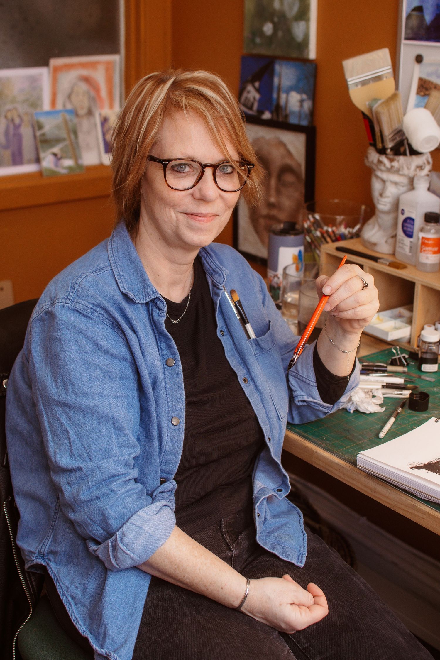 A smiling female artist with red hair and glasses holds a paintbrush in her art studio surrounded by paintings and supplies.