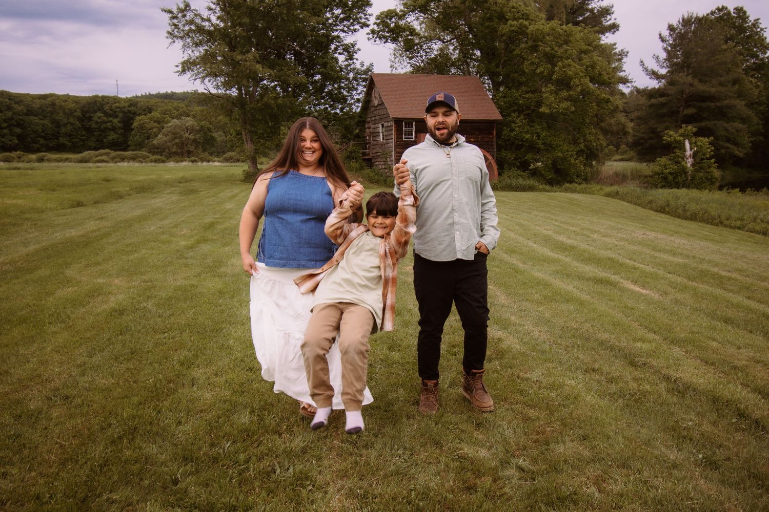 Family of three walking on green lawn, parents swinging child between them, brick house and trees in background.