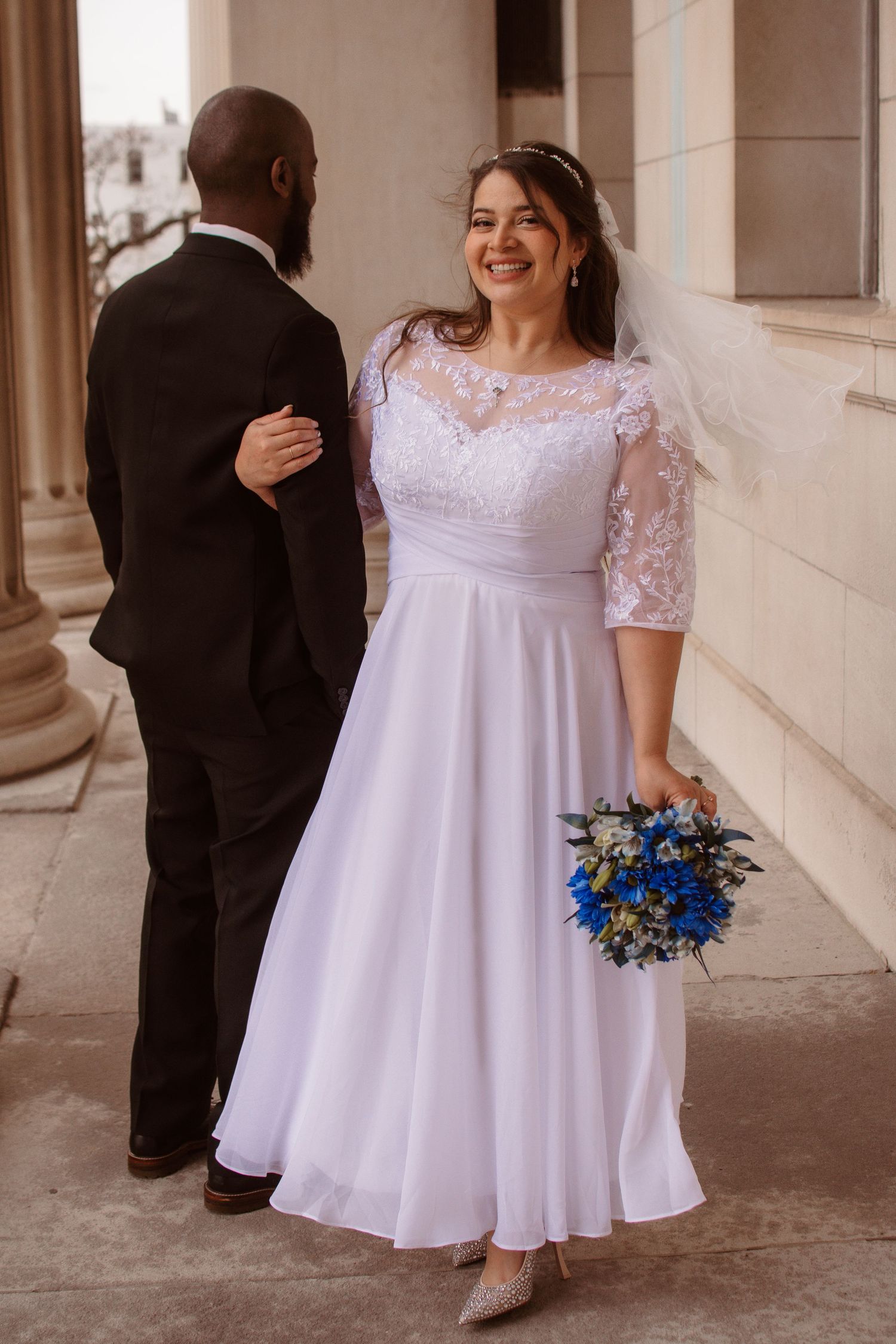 A wedding couple poses in a covered walkway with the bride wearing a white lace dress and holding a blue floral bouquet.