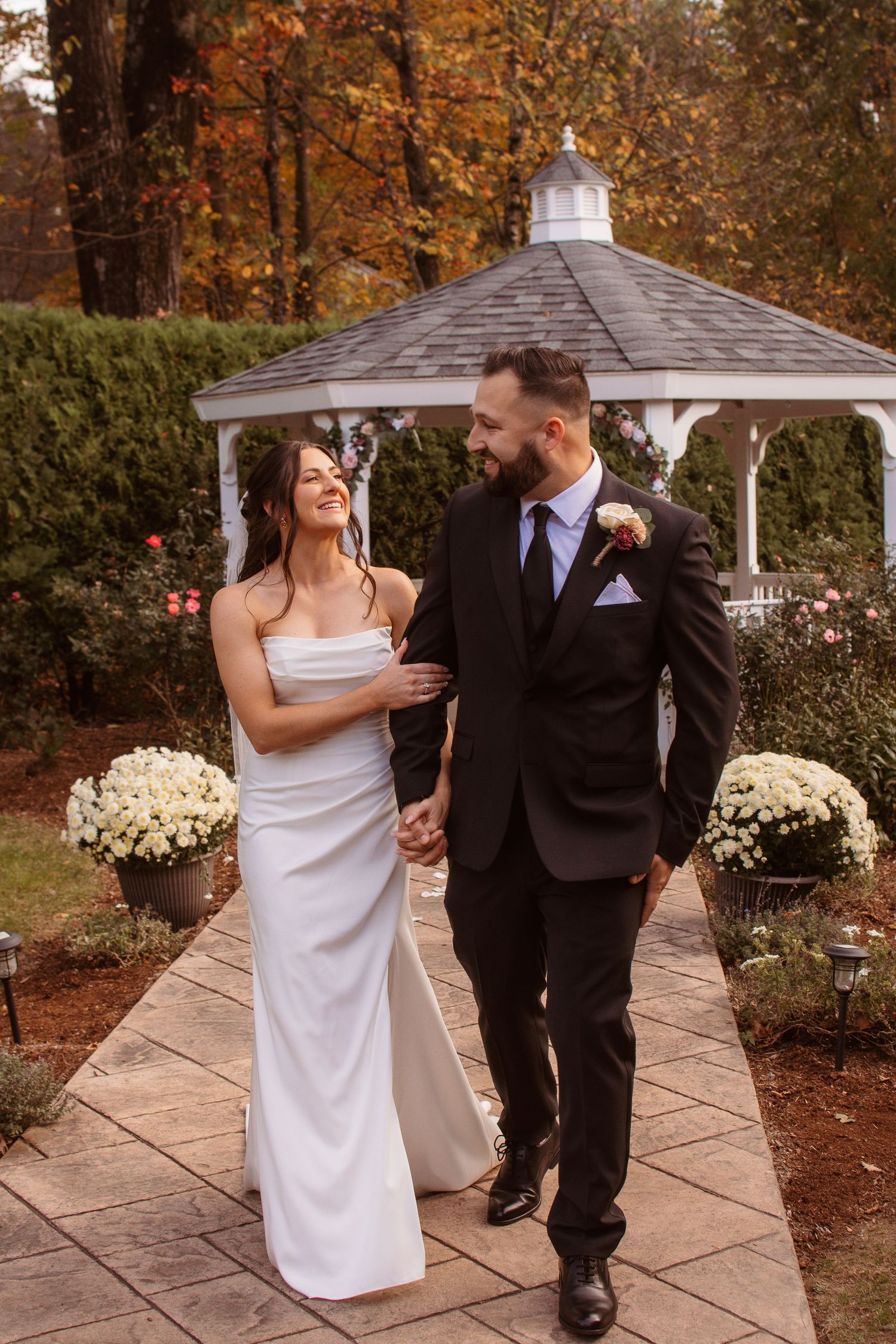 Wedding couple dancing and laughing together on stone patio with white gazebo and fall trees in background.