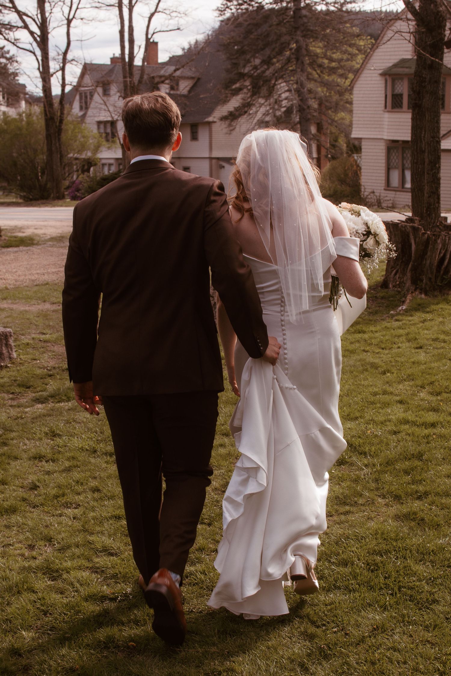 Bride and groom walking hand in hand away from camera, bride in white gown with veil, groom in dark suit, outdoor setting.