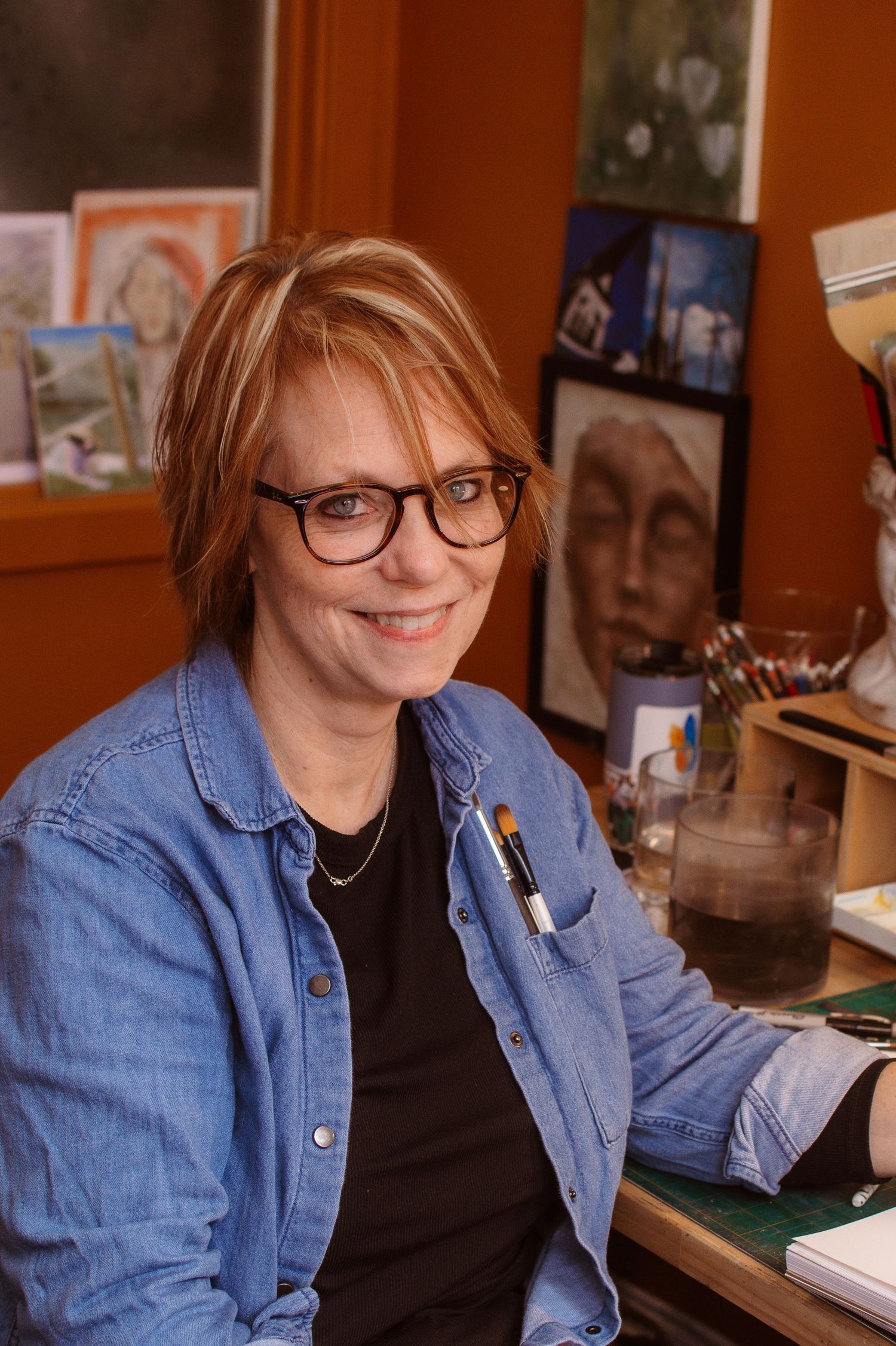 A smiling woman with glasses and short red hair sits at an artist's desk surrounded by paintings in her studio.
