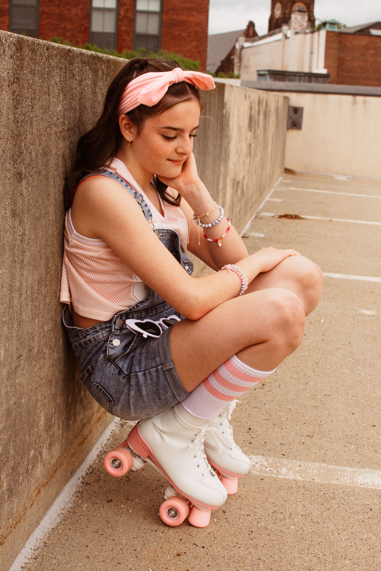 Someone in denim overalls and a pink top sitting casually against a concrete wall wearing vintage roller skates.