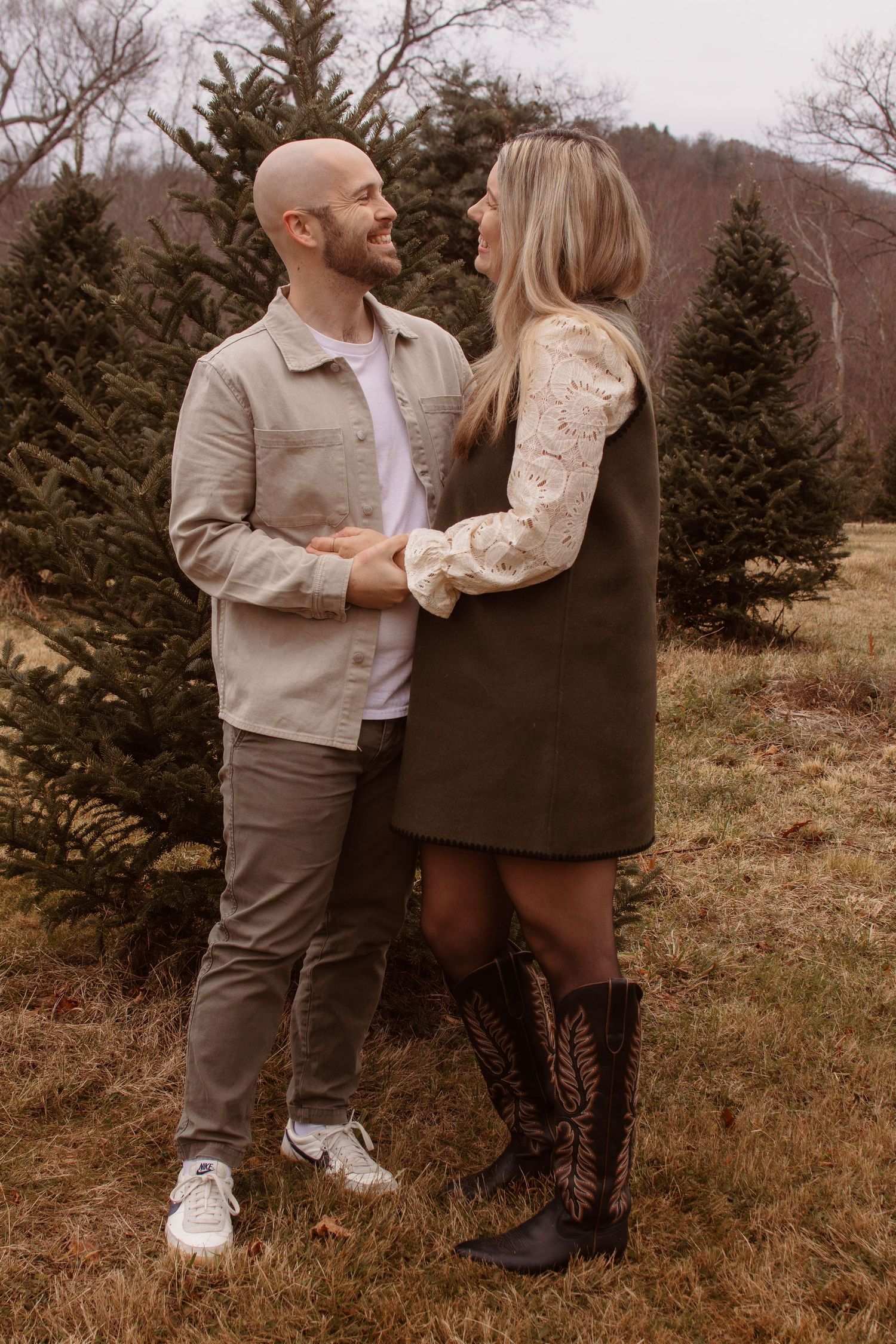 A couple embraces in a rustic outdoor setting with evergreen trees and brown winter foliage in the background.