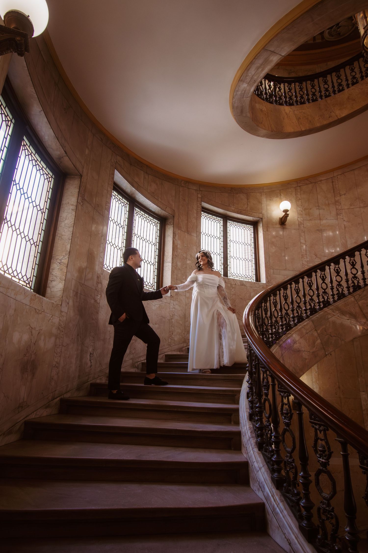Bride and groom holding hands on elegant curved staircase with ornate iron railings in a grand historic building.