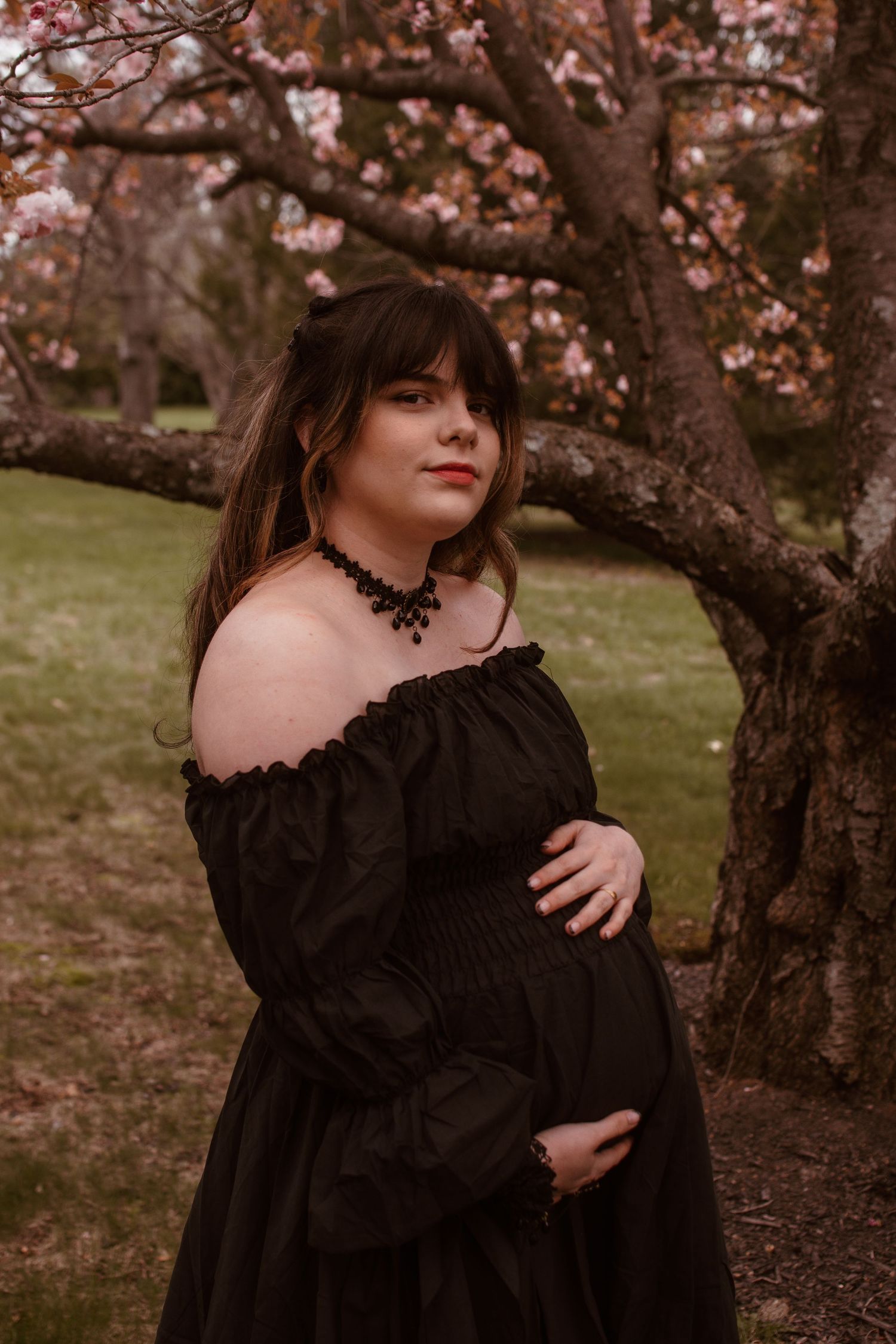 Pregnant woman in black off-shoulder gown poses under blooming cherry blossom trees during a maternity photoshoot.