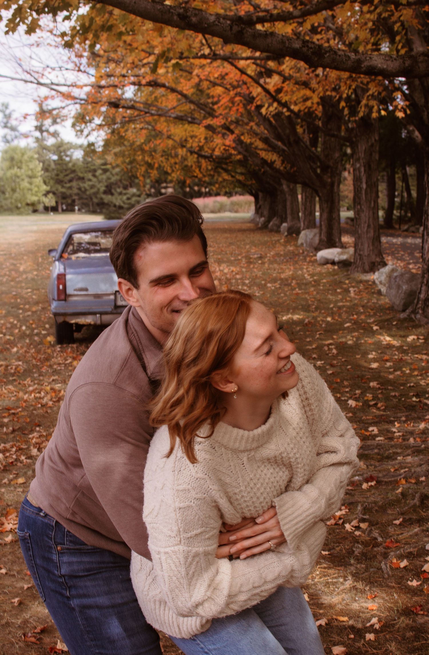 A couple shares a playful embrace under autumn trees with orange leaves falling in a park setting.