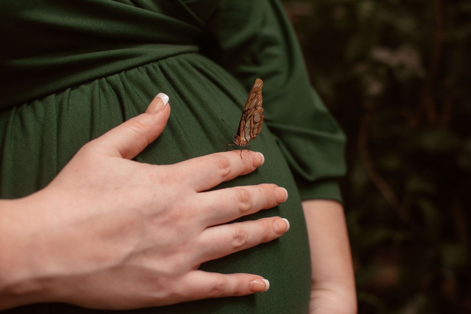 A delicate butterfly perches on a green dress fabric while a hand gently rests nearby in a nature setting.