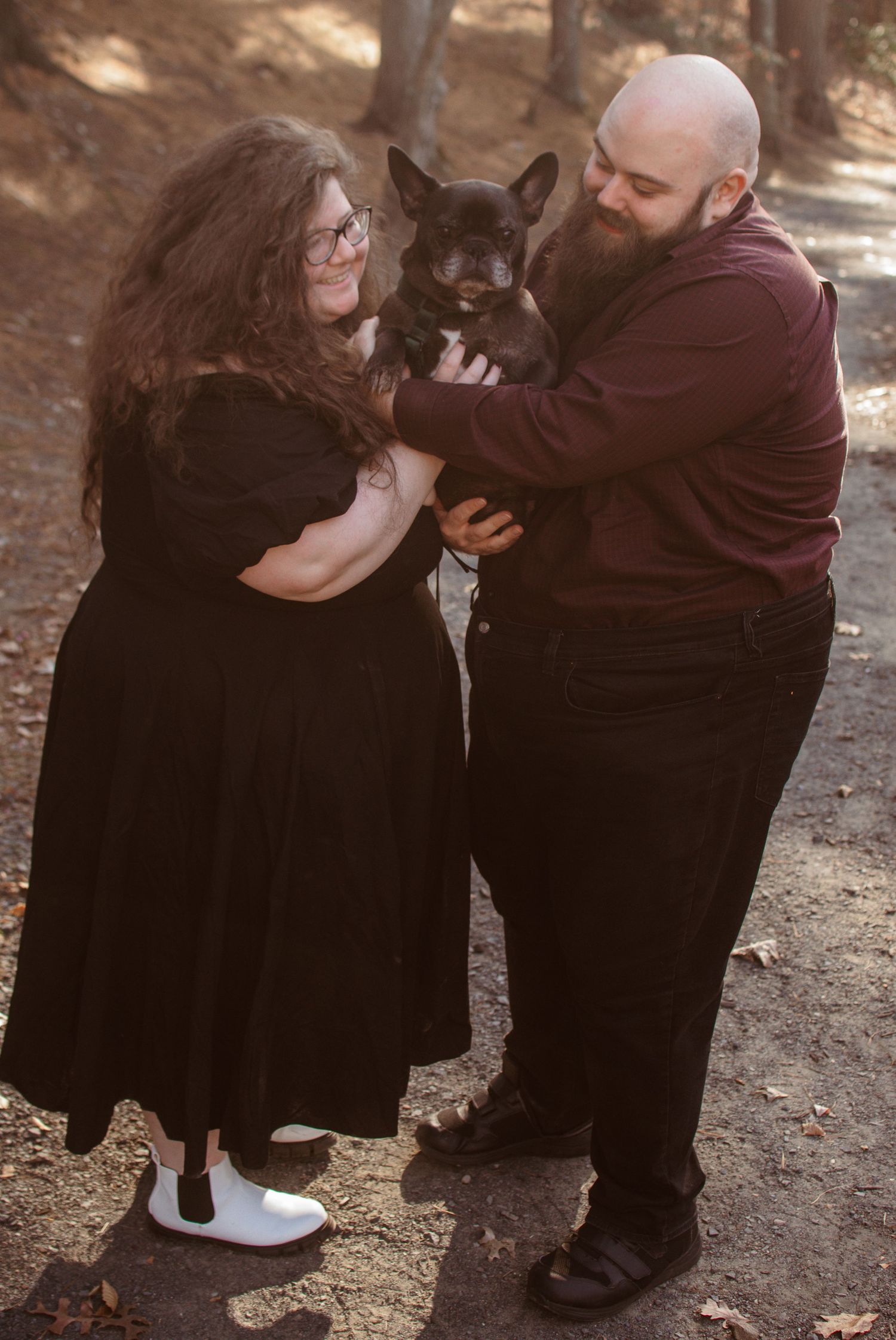 Couple in dark outfits dancing playfully and sharing an intimate moment on a sunlit dirt path through the trees.