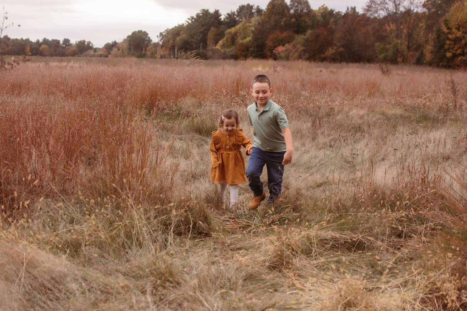 Two people walk hand in hand through a golden autumn field with tall grass and trees in the background.