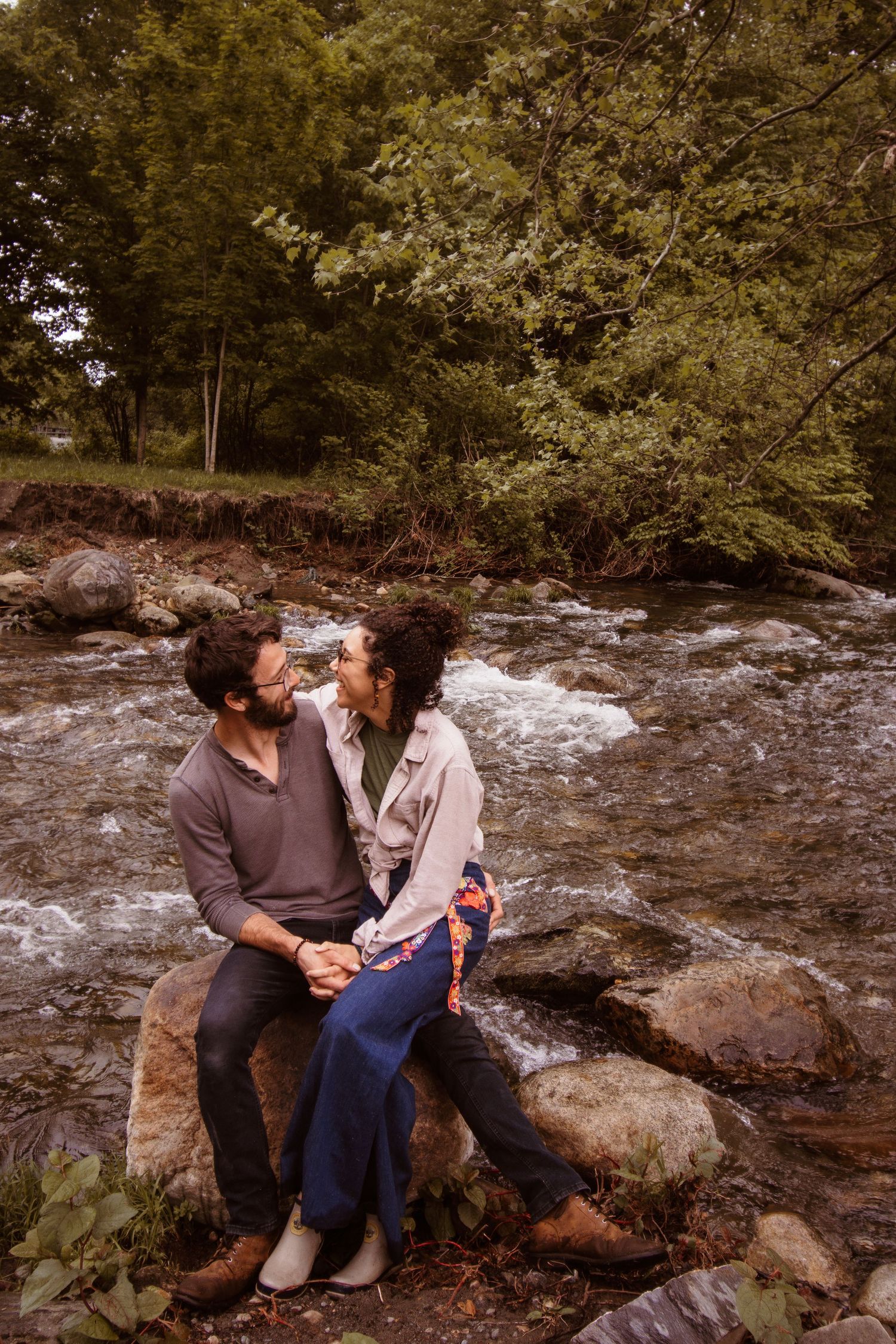 A couple sits together on rocks beside a rushing stream in the woods on an autumn day.