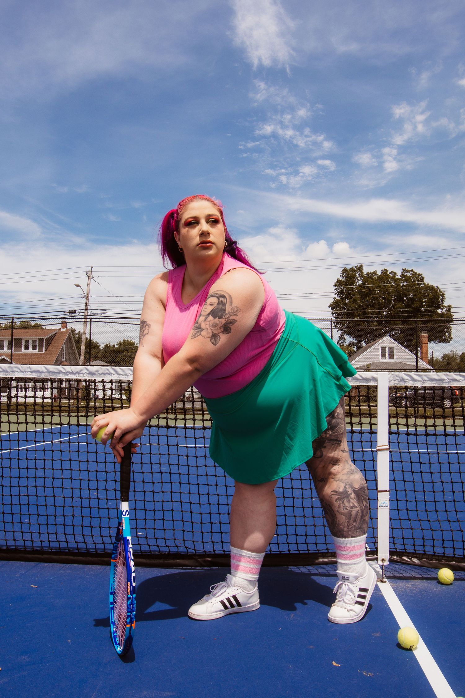 Tennis player in pink crop top and green skirt poses on blue court with racquet and visible tattoos.