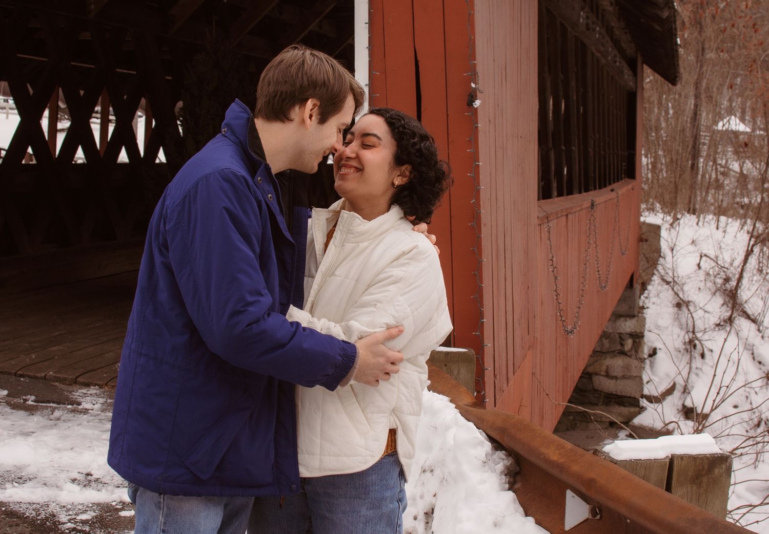 A couple in winter attire share a romantic moment in front of a rustic red barn door.