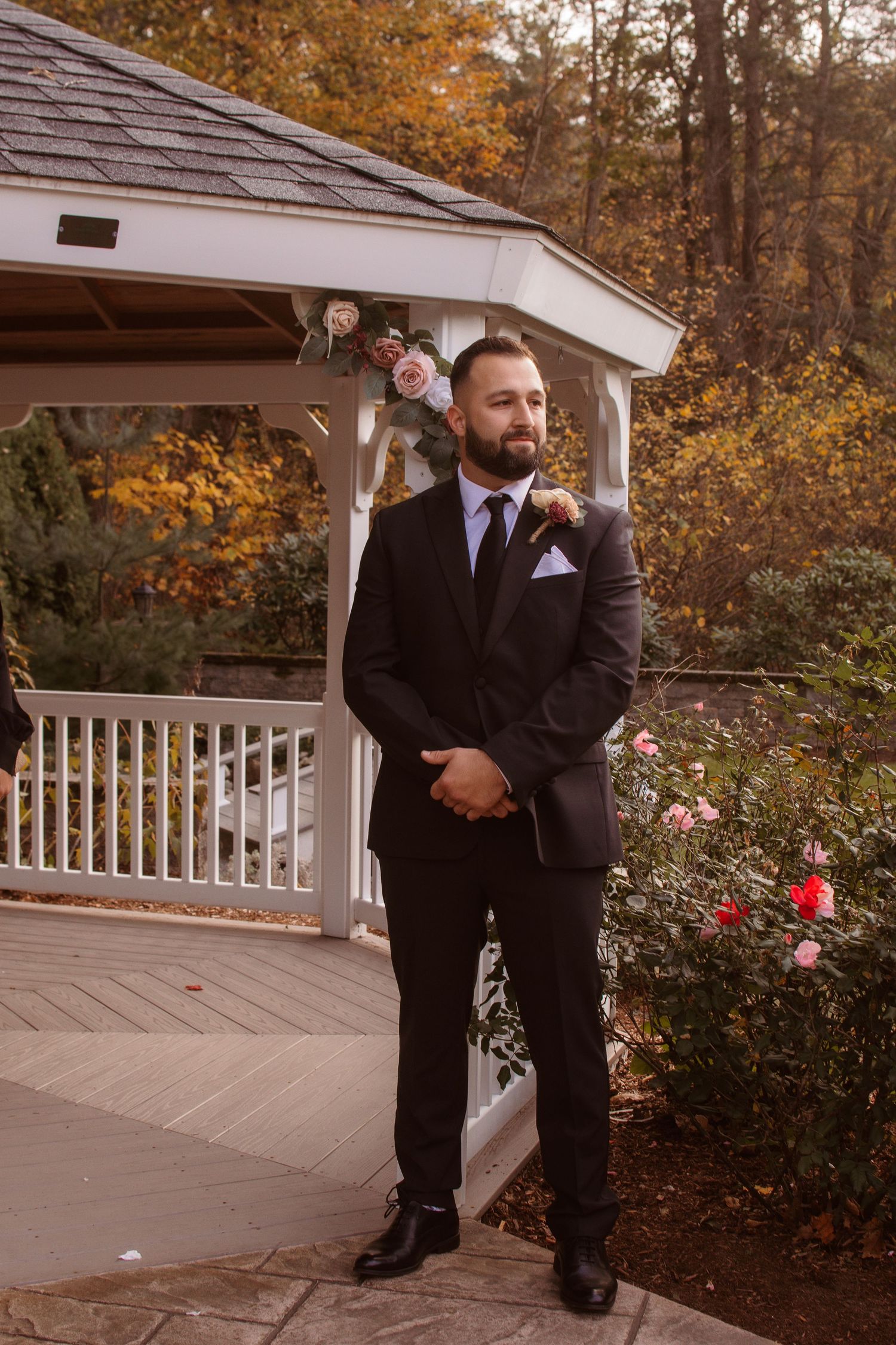 Groom in black tuxedo poses thoughtfully in autumn garden setting with white gazebo and fall foliage in background.
