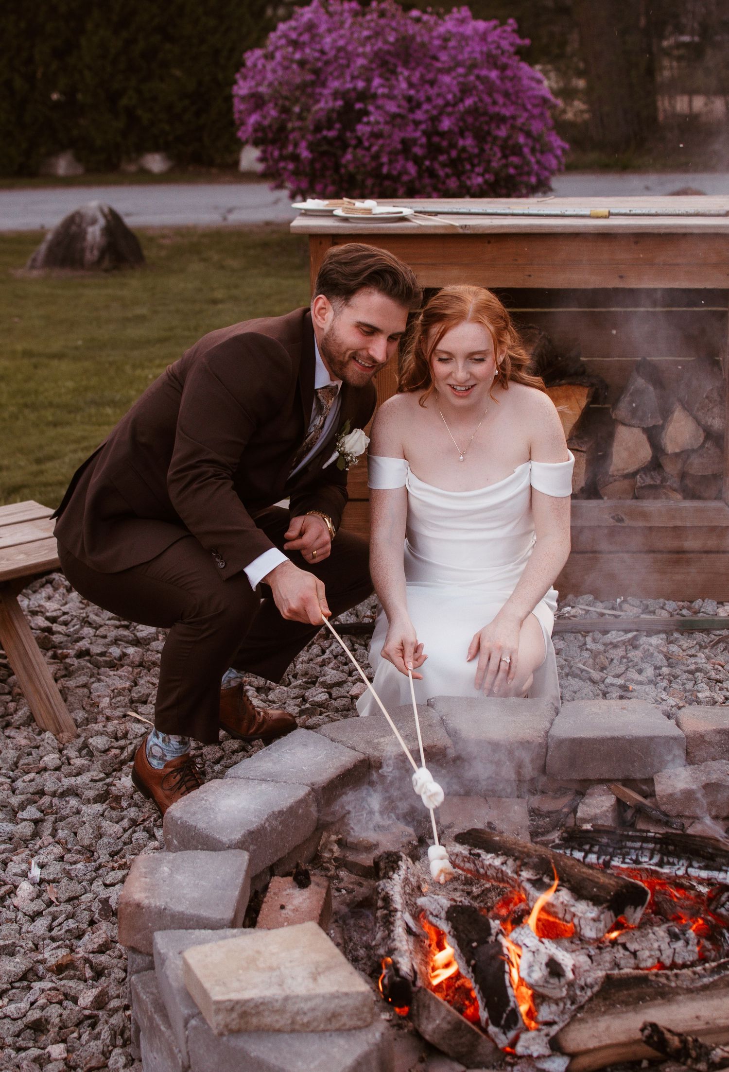Bride and groom roasting marshmallows over a fire pit at their outdoor wedding reception at dusk.