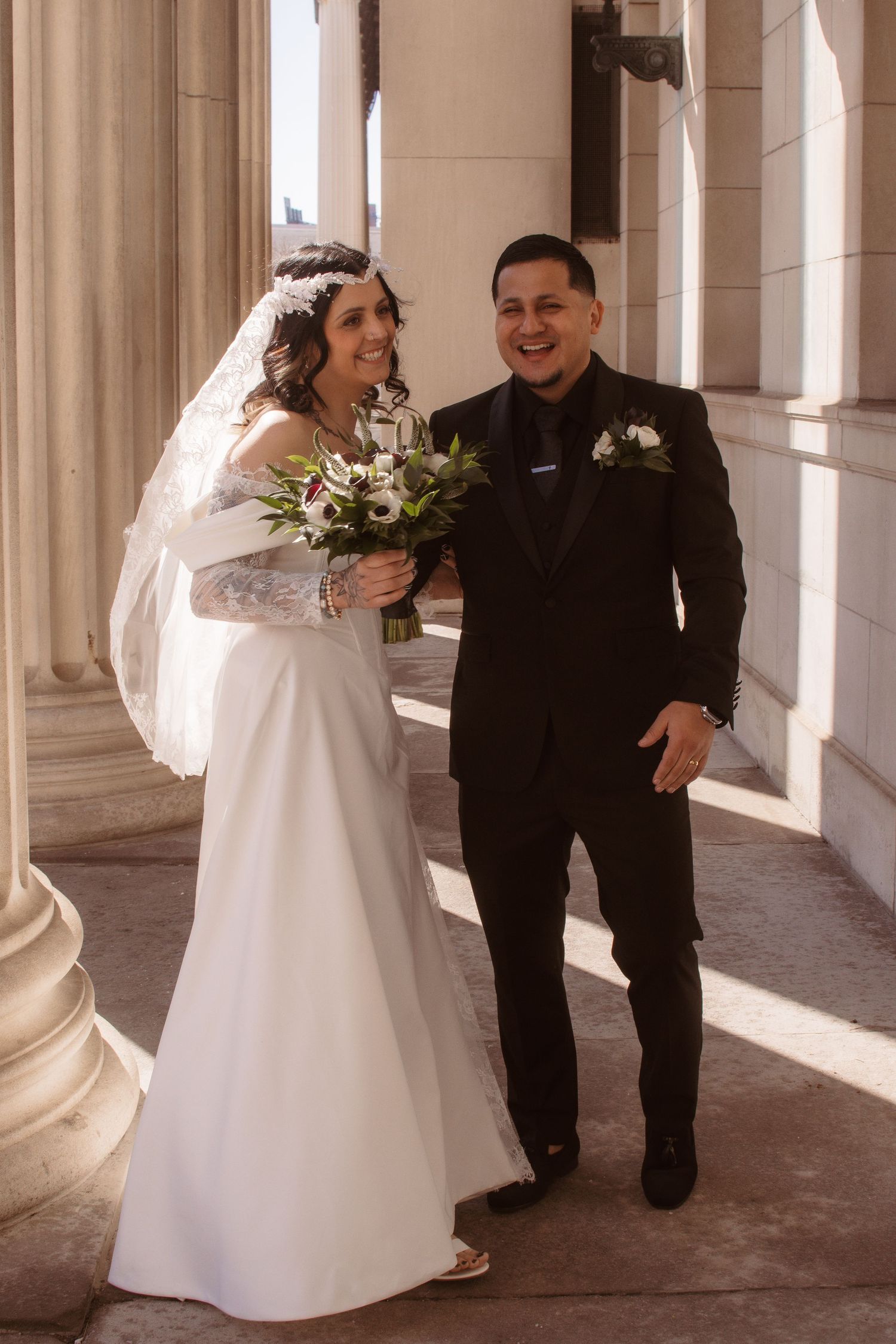 Smiling bride in white gown with veil and bouquet stands beside groom in black tuxedo with boutonniere by stone columns.