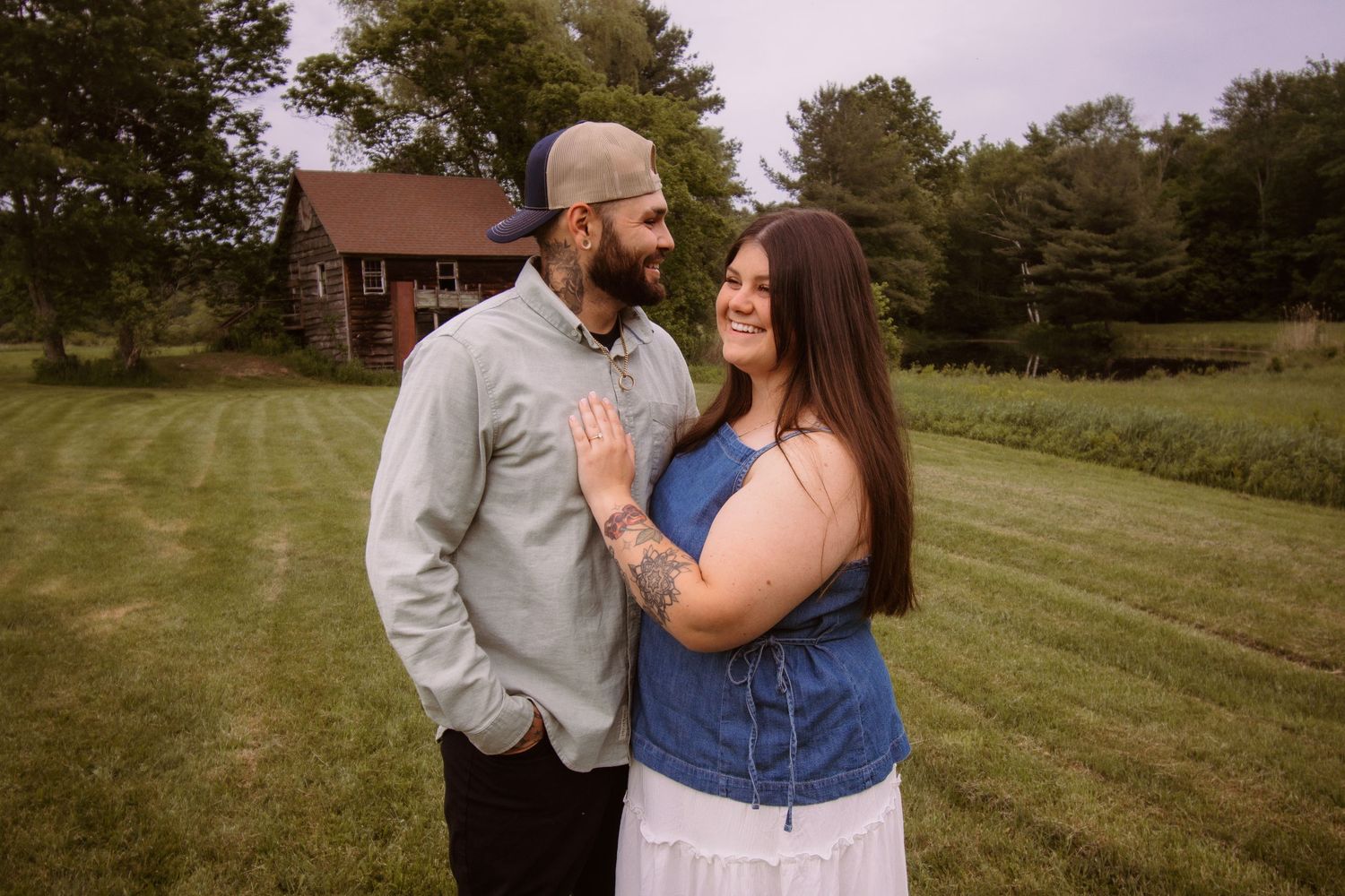 Couple smiling at each other in a green field with a red barn in the background during an outdoor engagement photo session.