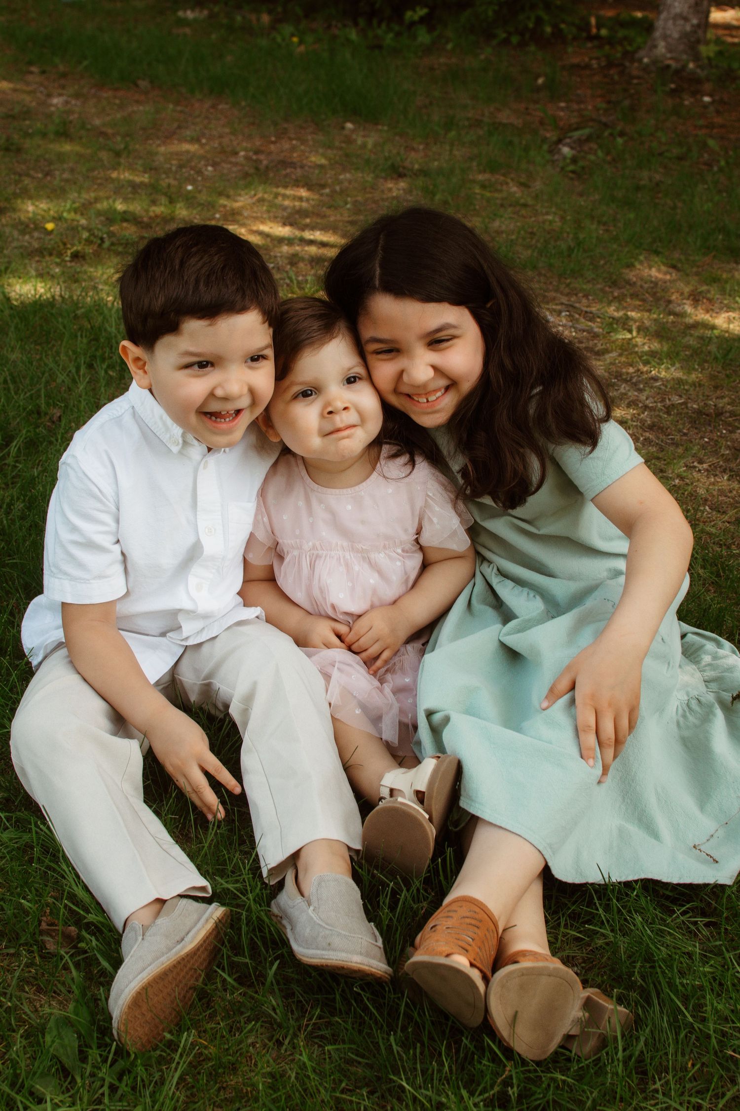 Children sit together outdoors on grassy ground wearing coordinated pastel outfits in natural afternoon light.