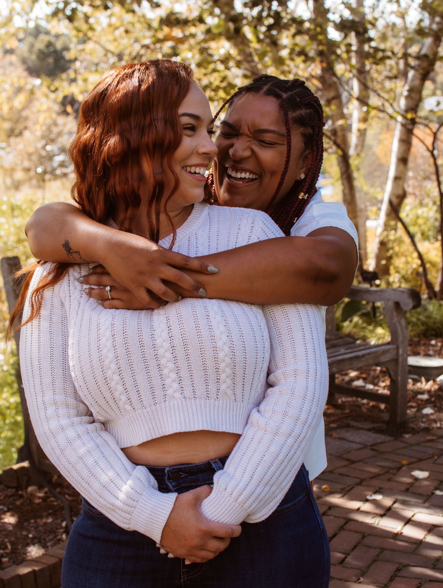 Two people in white sweaters share warm embraces and laughter in a sunny outdoor setting with brick and foliage.