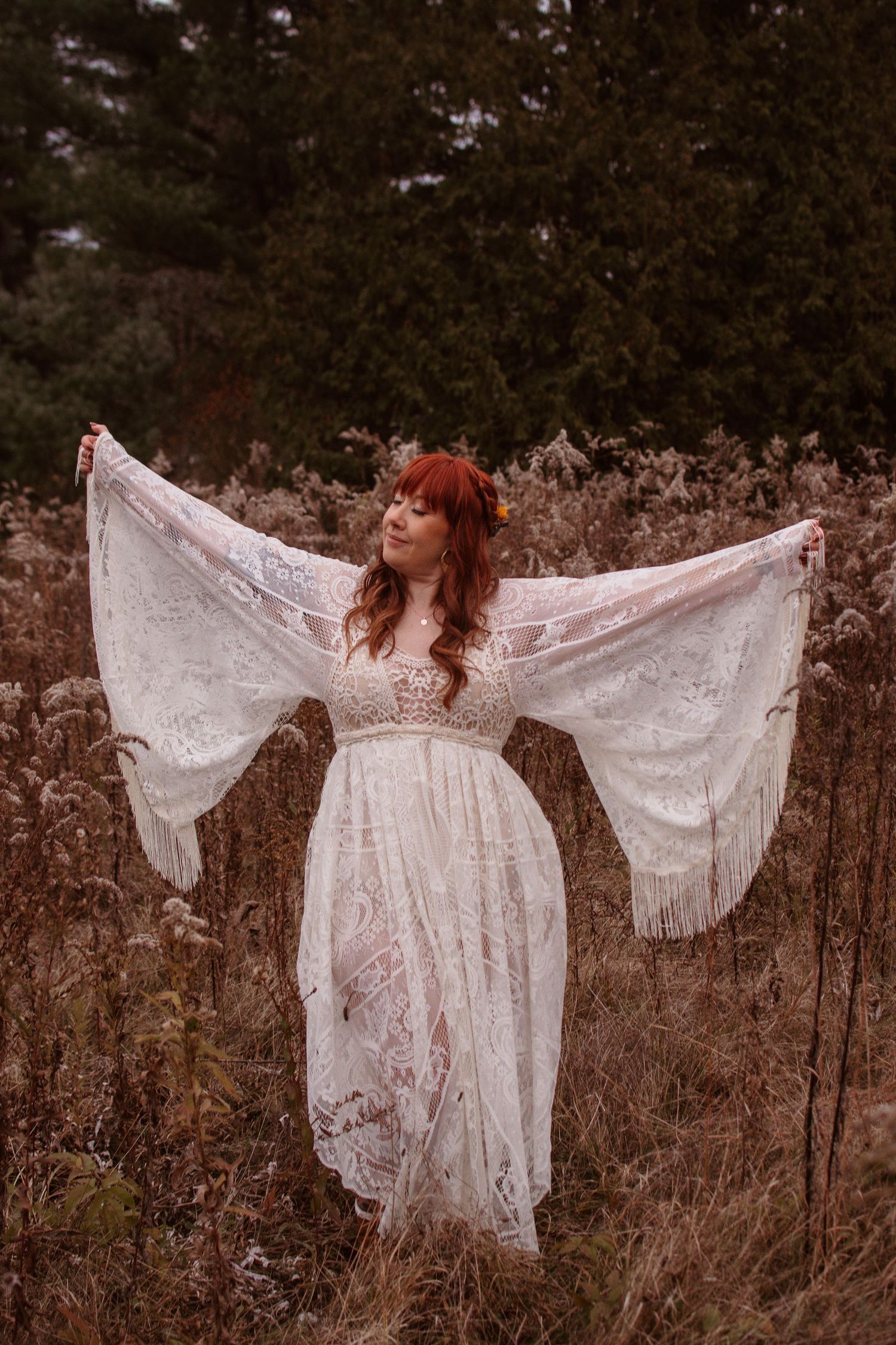 A figure in a flowing white lace dress with bell sleeves stands gracefully in a field of dried grass and evergreens.