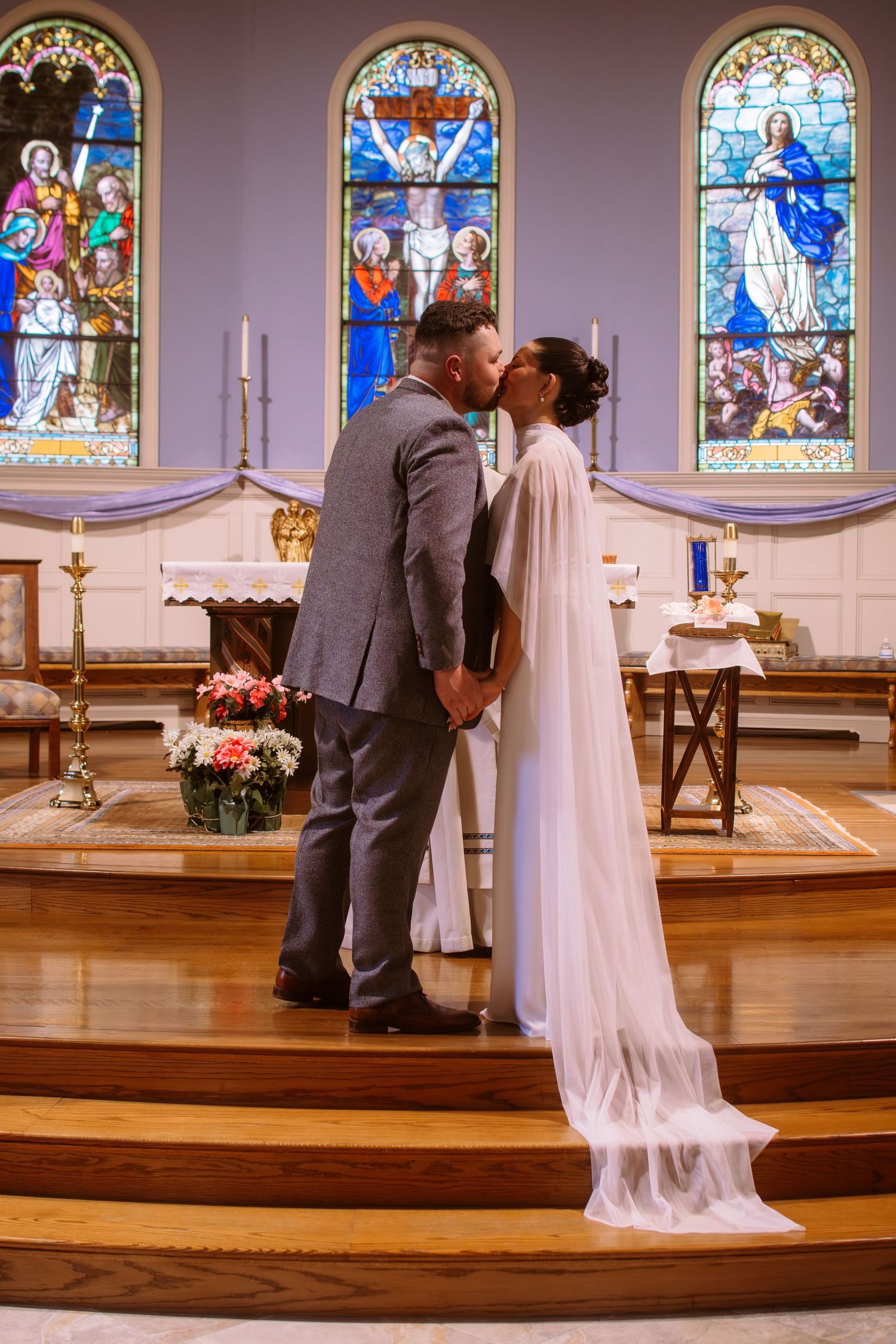 Newlywed couple sharing first kiss at church altar with colorful stained glass windows behind them.
