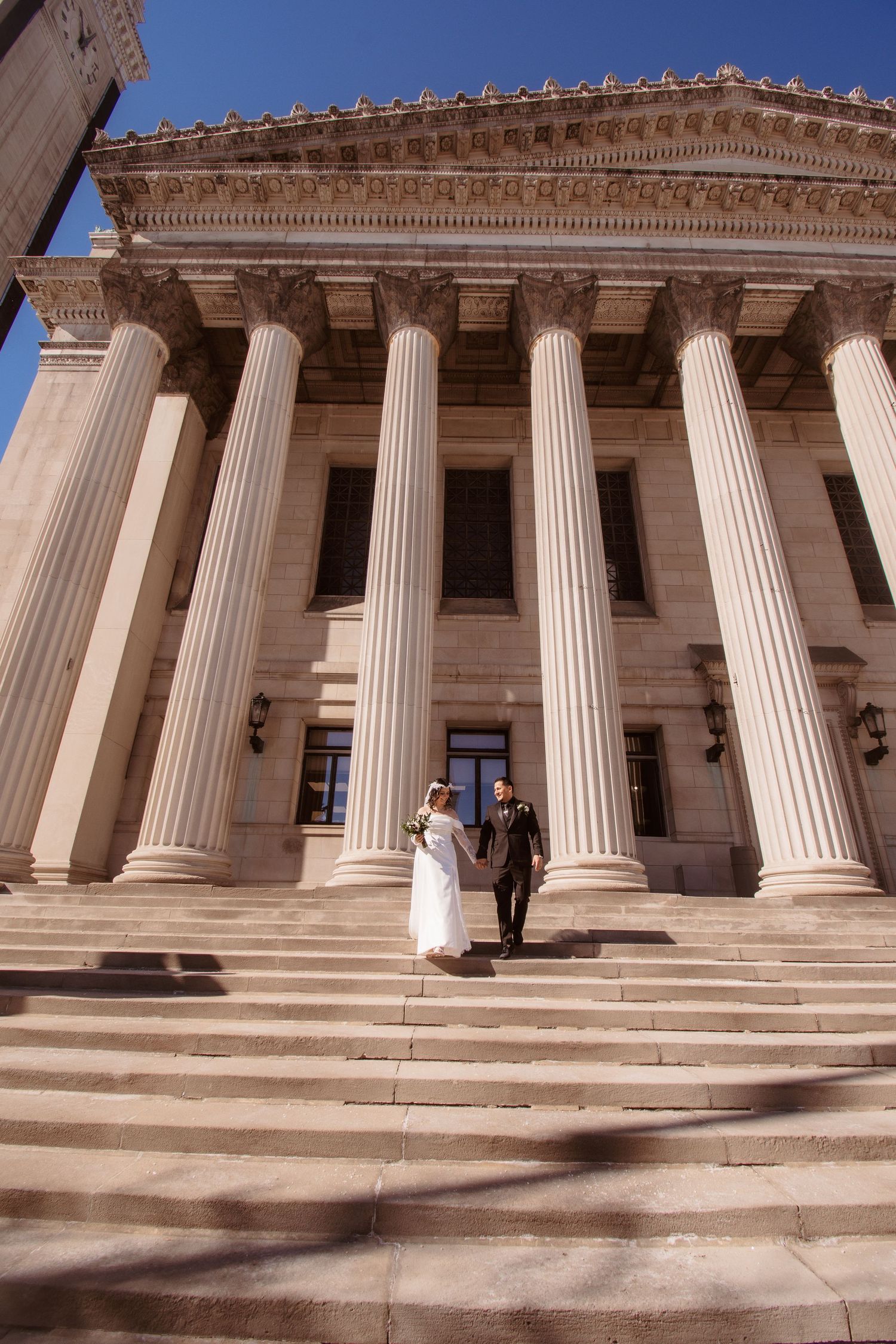 Bride and groom walking down grand courthouse steps with tall columns on their wedding day.