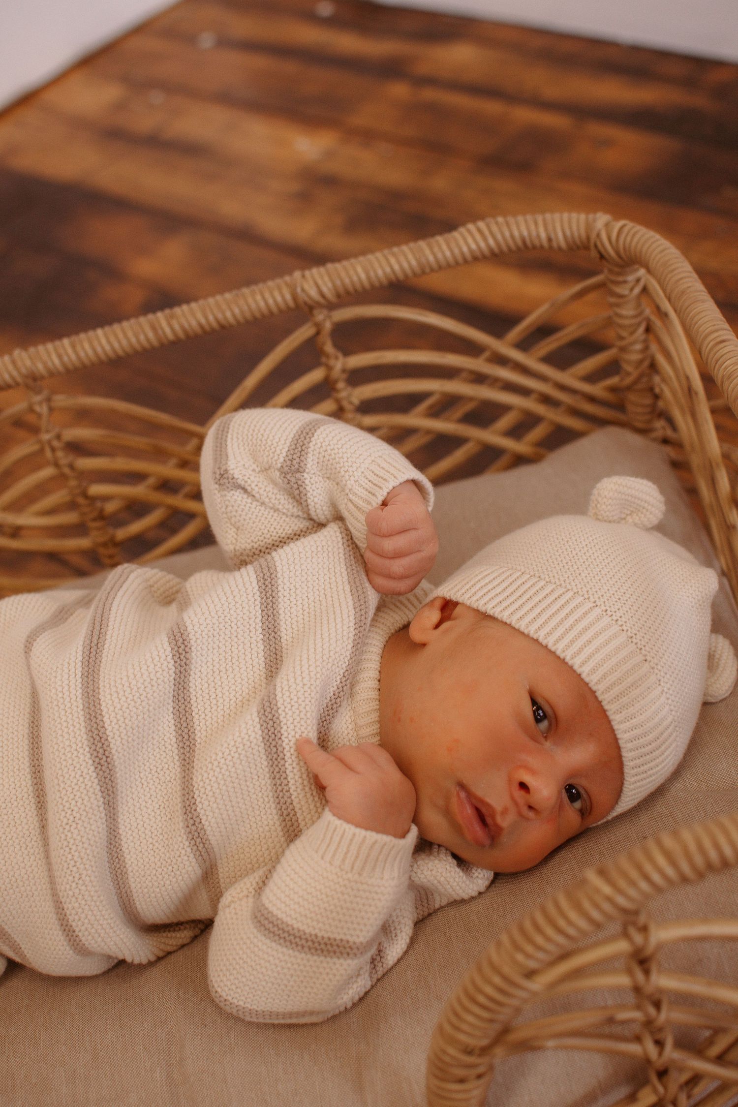 Newborn baby in cream striped knit outfit and bear ear hat lying in a wicker bassinet.