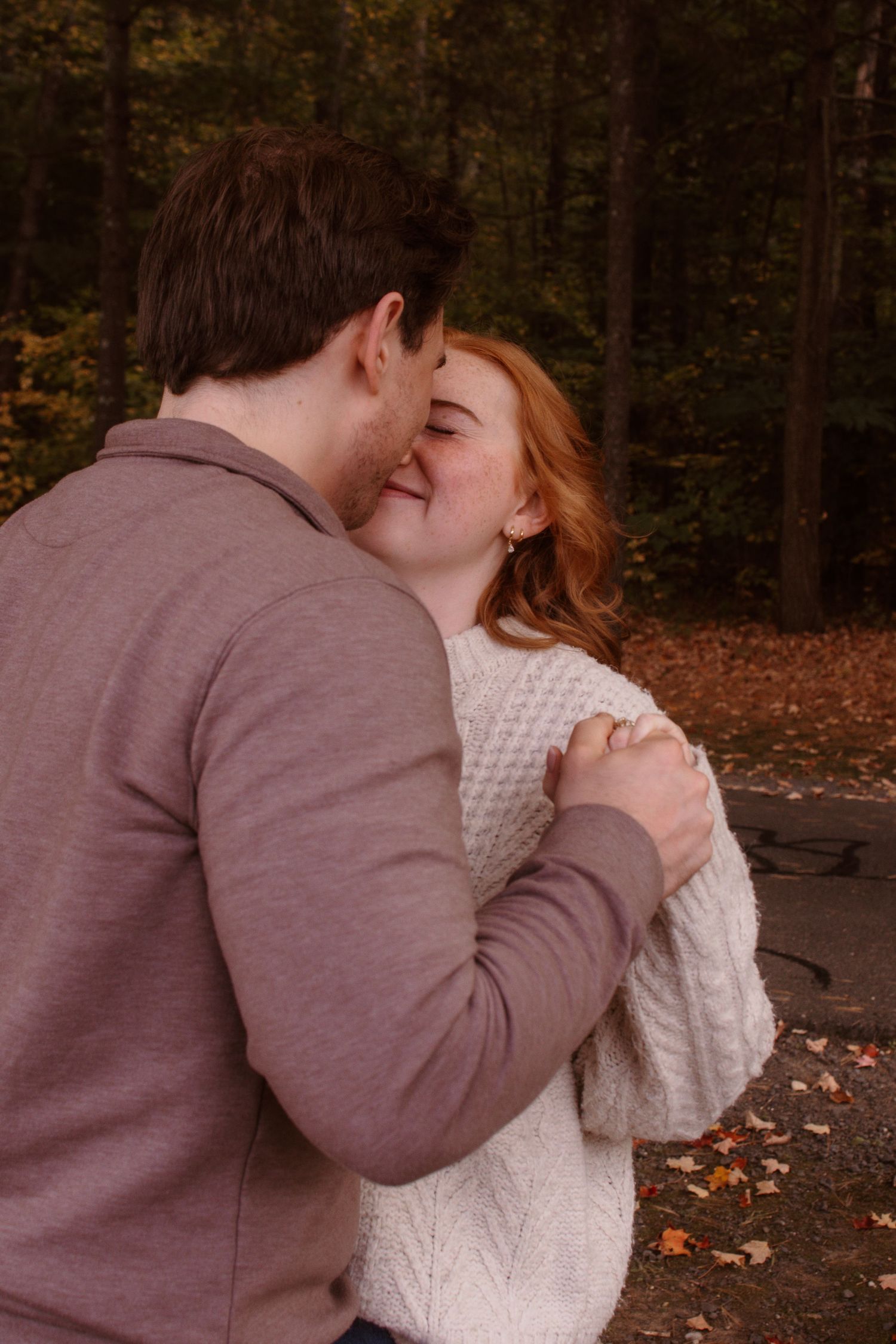 A couple in autumn woods share an intimate embrace with person in brown sweater and white knit sweater.
