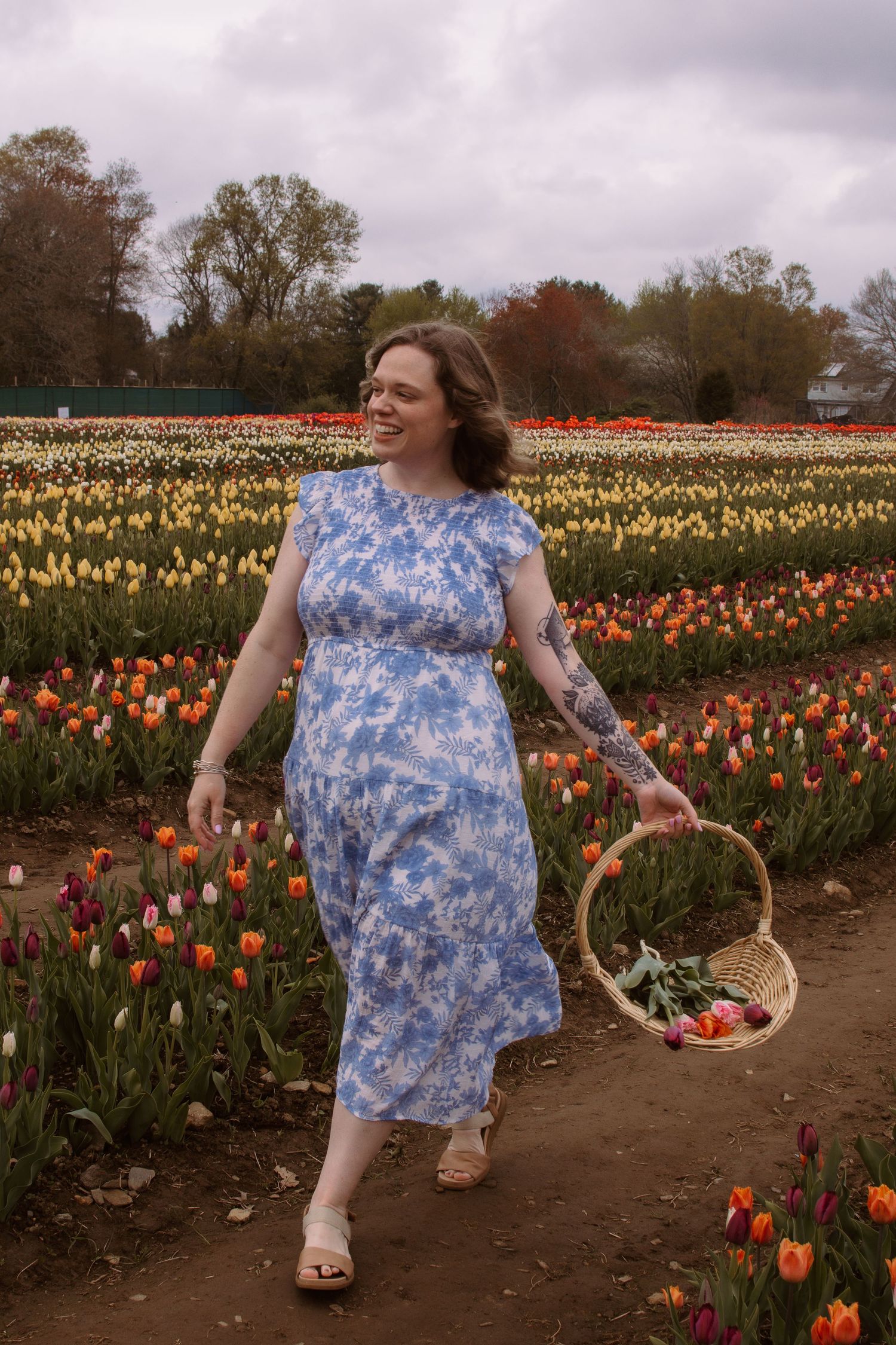 Woman in blue floral dress carrying a wicker basket walks through a colorful tulip field.