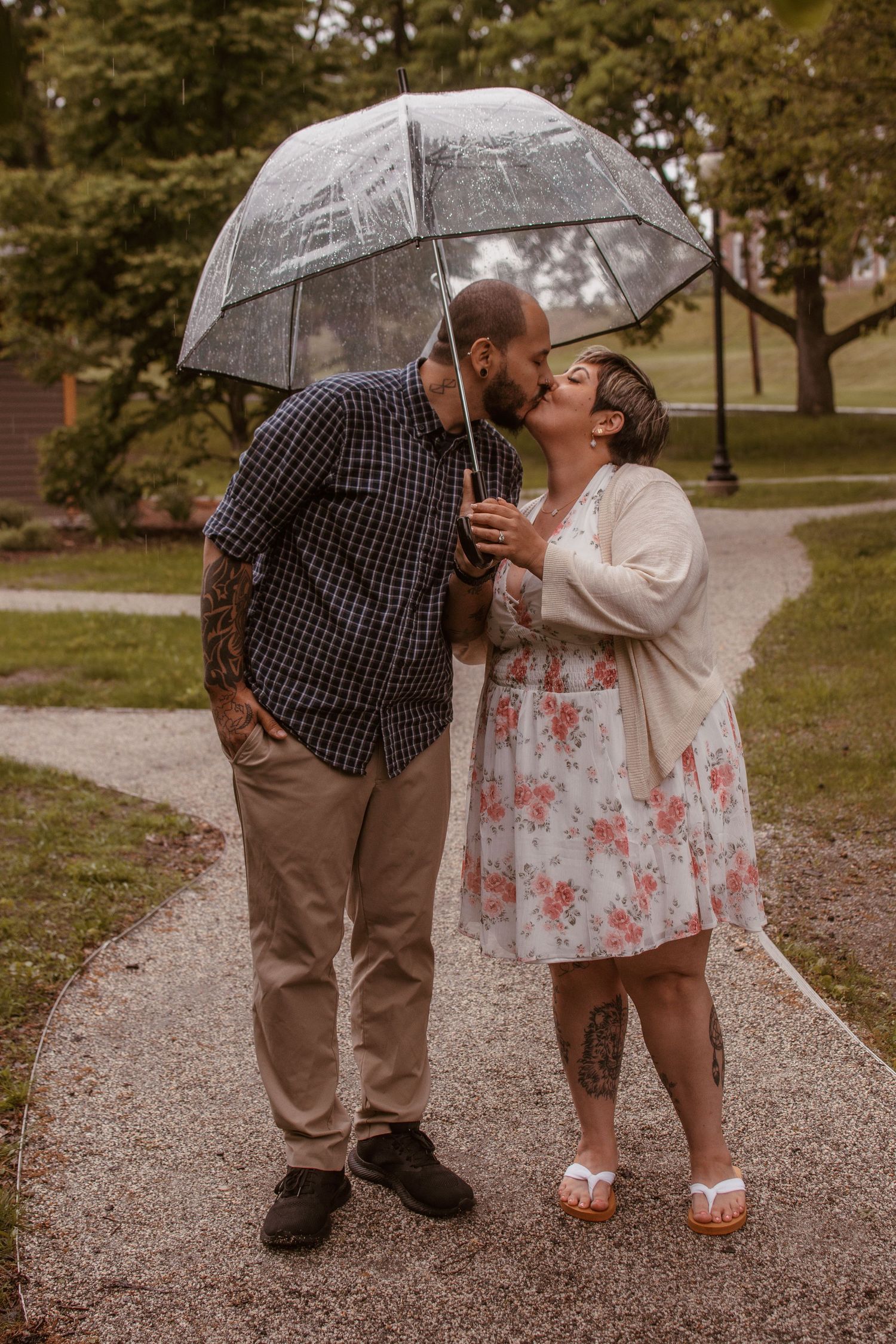 Couple kissing under a clear bubble umbrella on a rainy park path, man with tattoos and woman in floral dress.